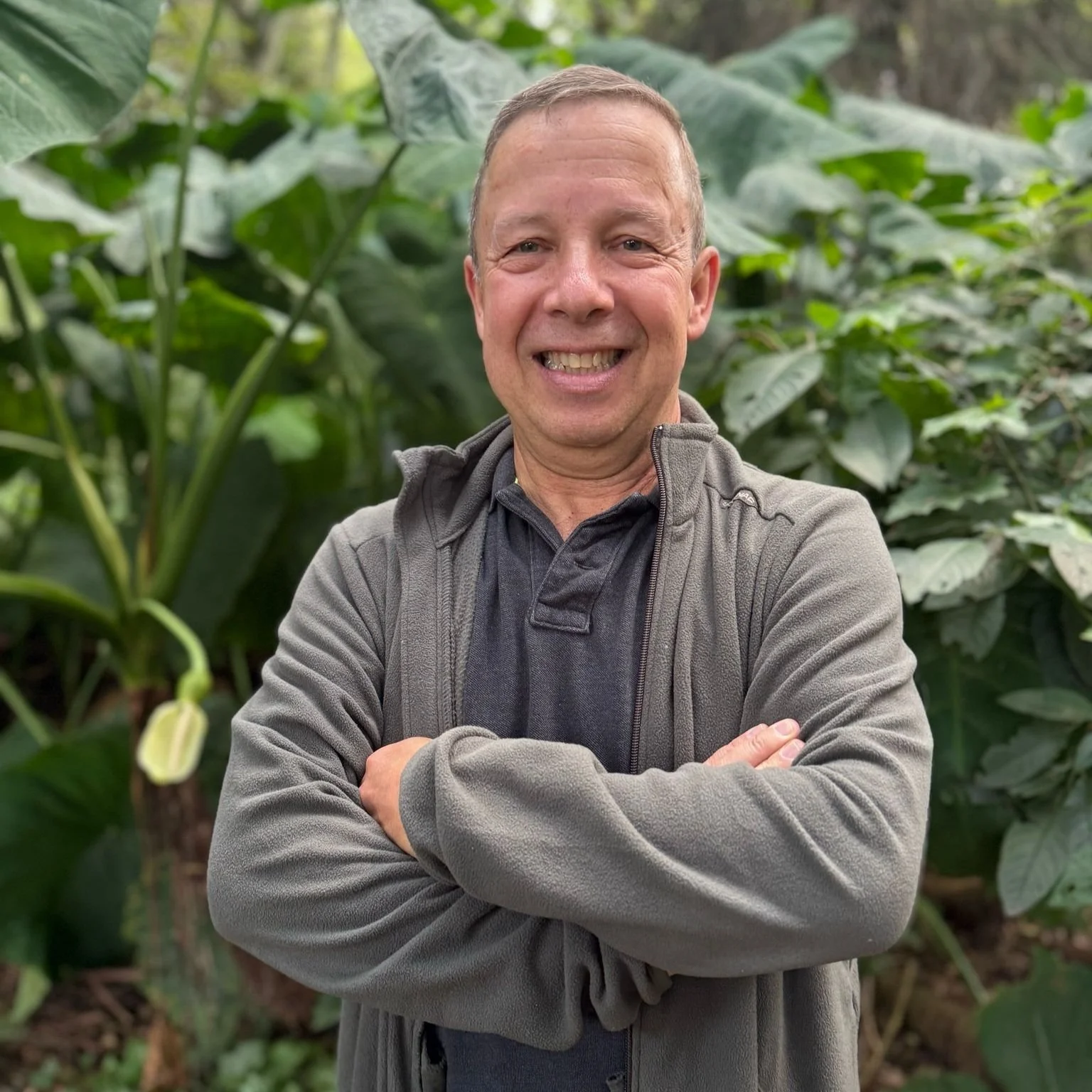 Hombre sonriendo con brazos cruzados, de pie frente a plantas verdes.