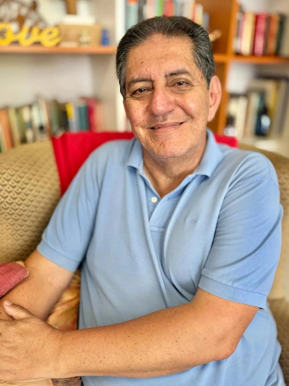 Hombre sonriendo sentado en una sala con libros de fondo.