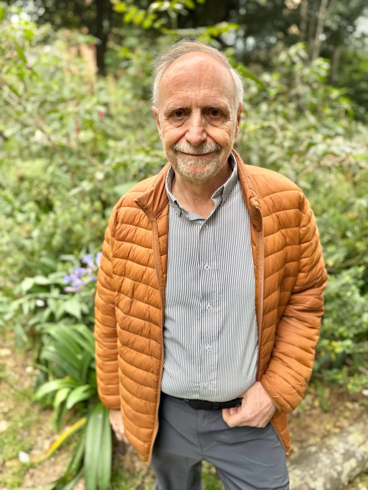 Hombre mayor con barba y camiseta de rayas, usando chaqueta marrón, en un entorno natural con vegetación