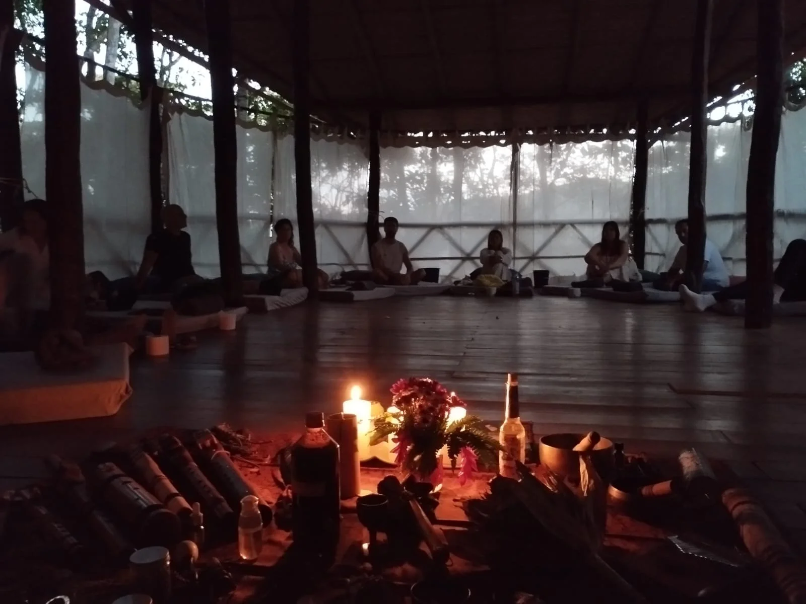 Grupo de personas en círculo en un espacio de madera, con velas y objetos en el centro, durante la noche o al atardecer.