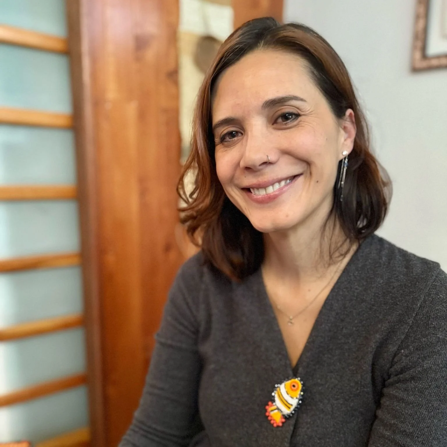 Mujer con cabello castaño y cabello rizado, sonriendo, usando un arete largo y un collar con un pez de colores, en un entorno interior con paredes de madera y cuadros en el fondo.