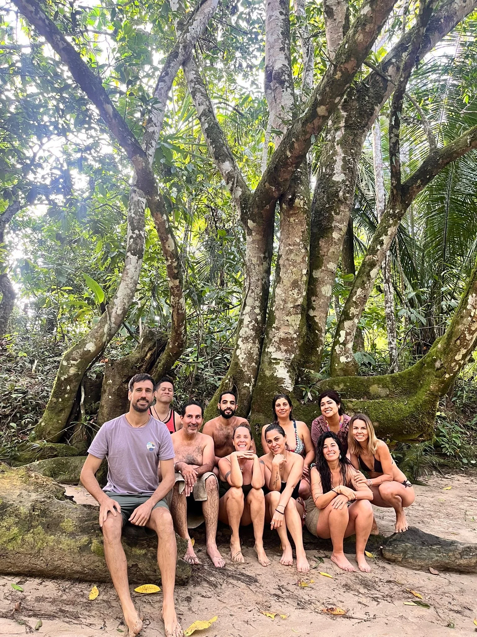 Grupo de personas en un bosque tropical, sentados y de pie, con árboles grandes y vegetación densa alrededor.