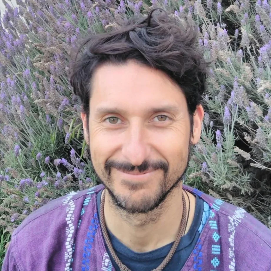 Hombre con cabello rizado y barba, sonriendo, frente a un campo de flores moradas y verdes.