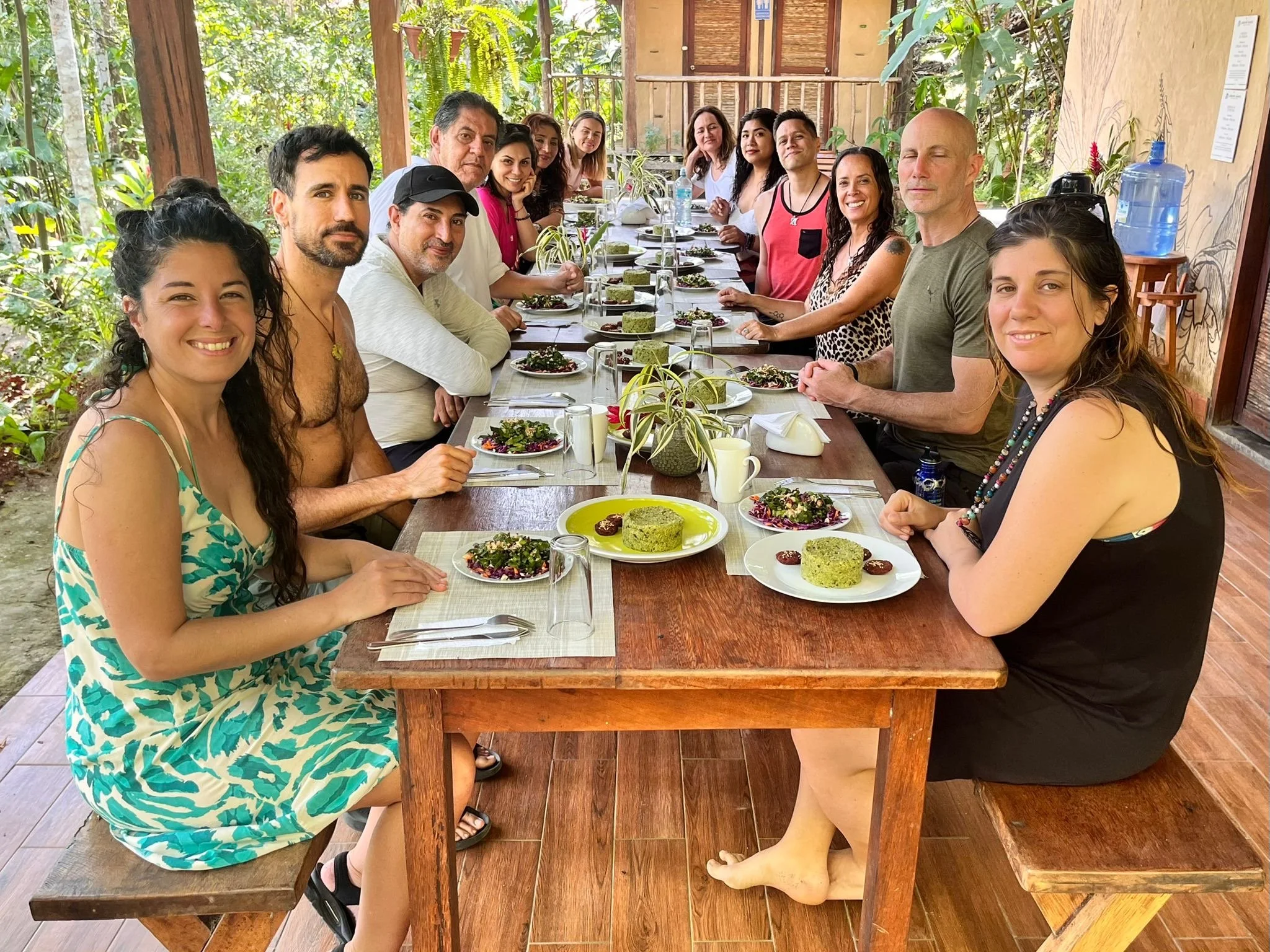 Grupo de personas sentadas en una mesa de madera en un restaurante abierto rodeado de vegetación, disfrutando de una comida con platos decorativos y bebidas.