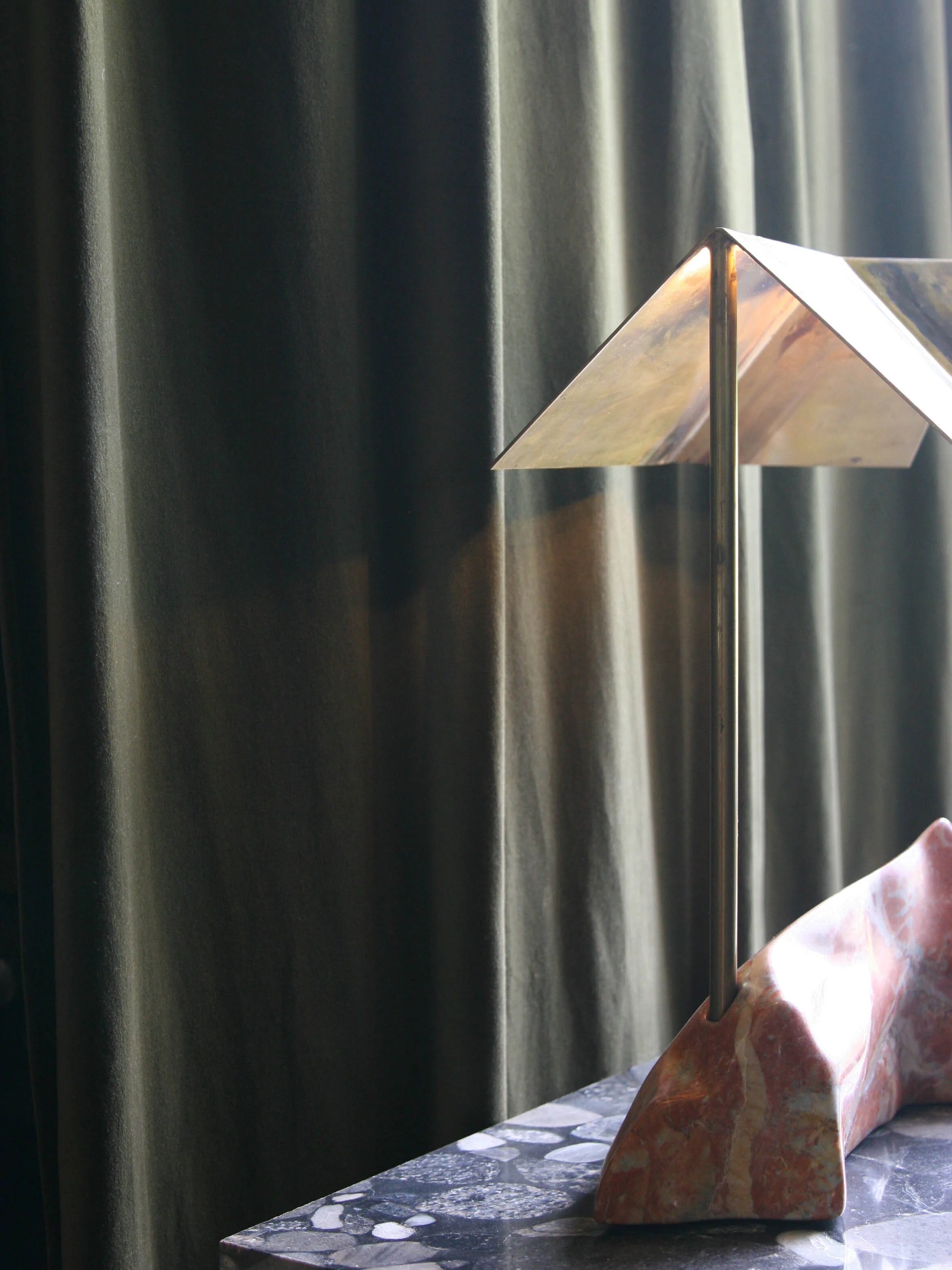 Close-up of a modern sculpture with a polished metal angled top, a thin vertical rod, and a multicolored stone base, set against a dark curtain background.