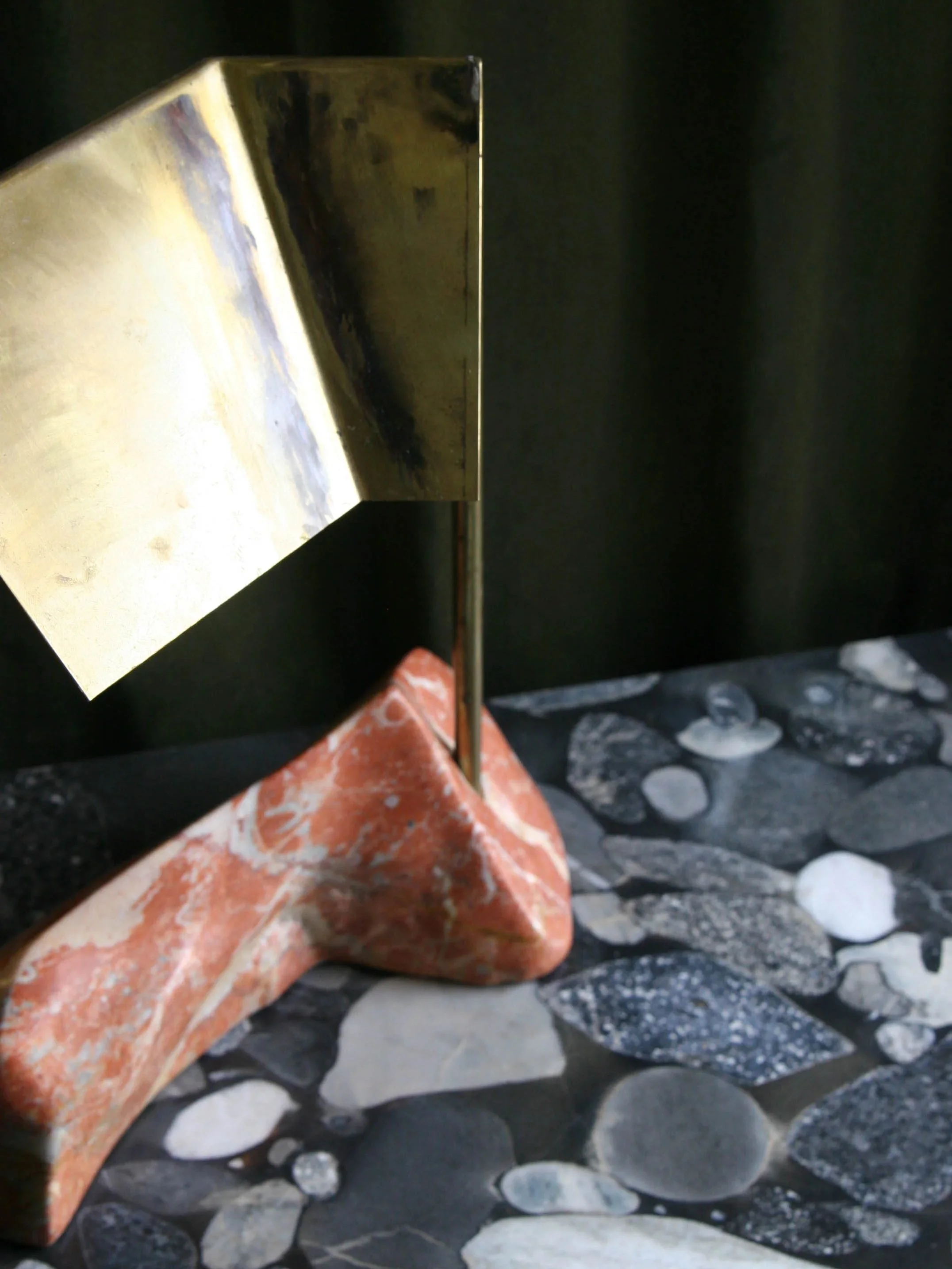 Modern decorative sculpture composed of a gold-colored metal structure supported on an irregularly shaped reddish stone, with a background of dark curtains and a pebble-patterned surface.