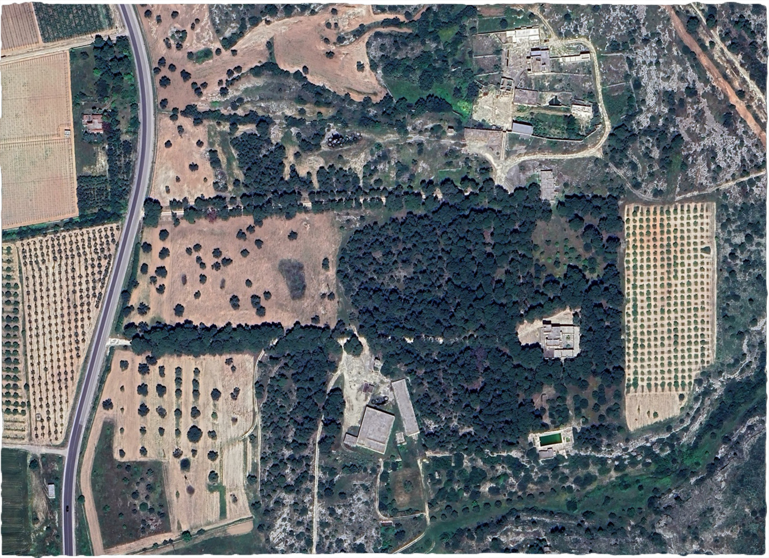 Satellite view of a semi arid landscape of creams, browns, greens and grays with winding and straights paths, orchards, forests, fields, buildings and ruins and a pool.