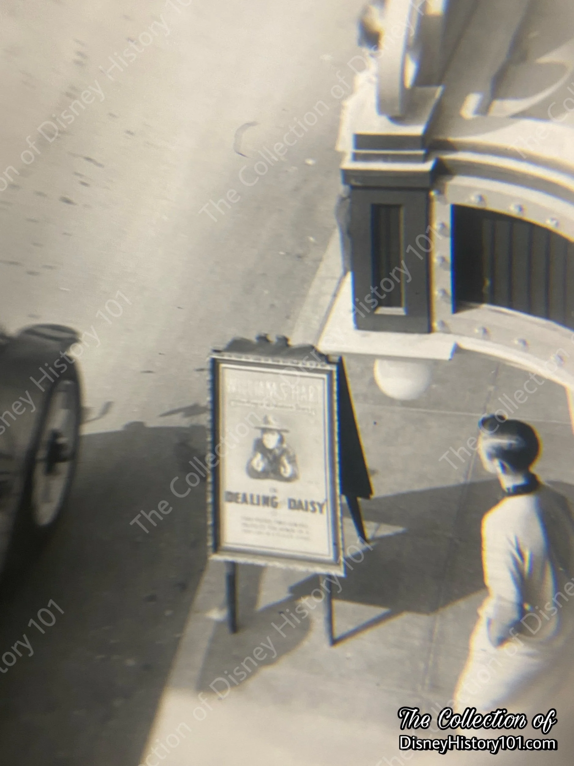 Through the loupe we can spy an A-frame “sandwich board” sidewalk sign promoting “A Dealing for Daisy” (1915) starring William S. Hart.