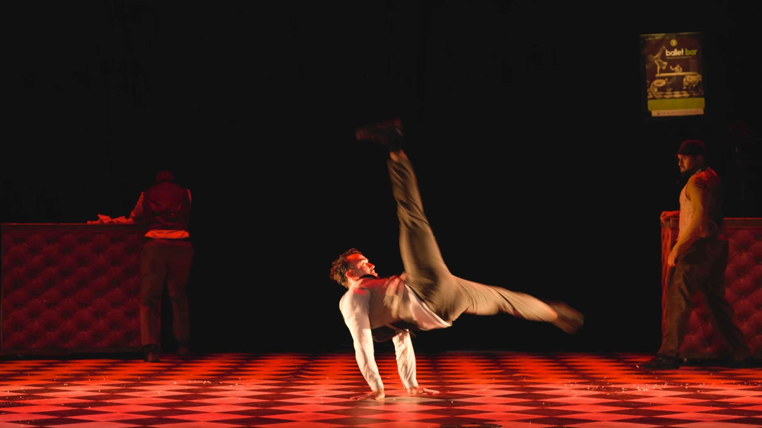 Un performeur breakdancer effectue un mouvement sur le sol dans un spectacle sur scène, avec deux autres personnes en arrière-plan dans un décor sombre et éclairages rouges.