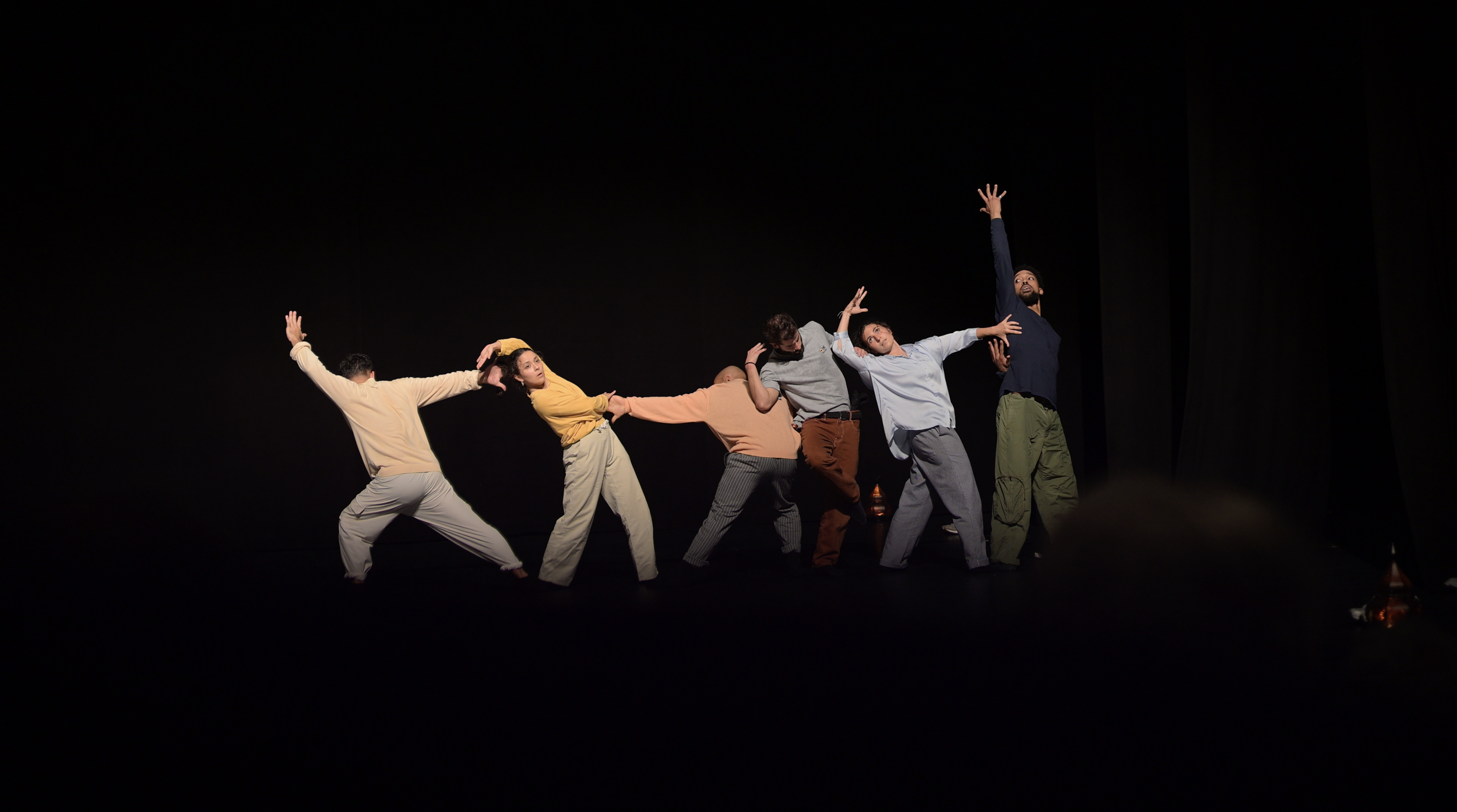 Groupe de sept personnes en train de danser sur une scène sombre, en position expressive avec des mouvements de bras et de corps variés.