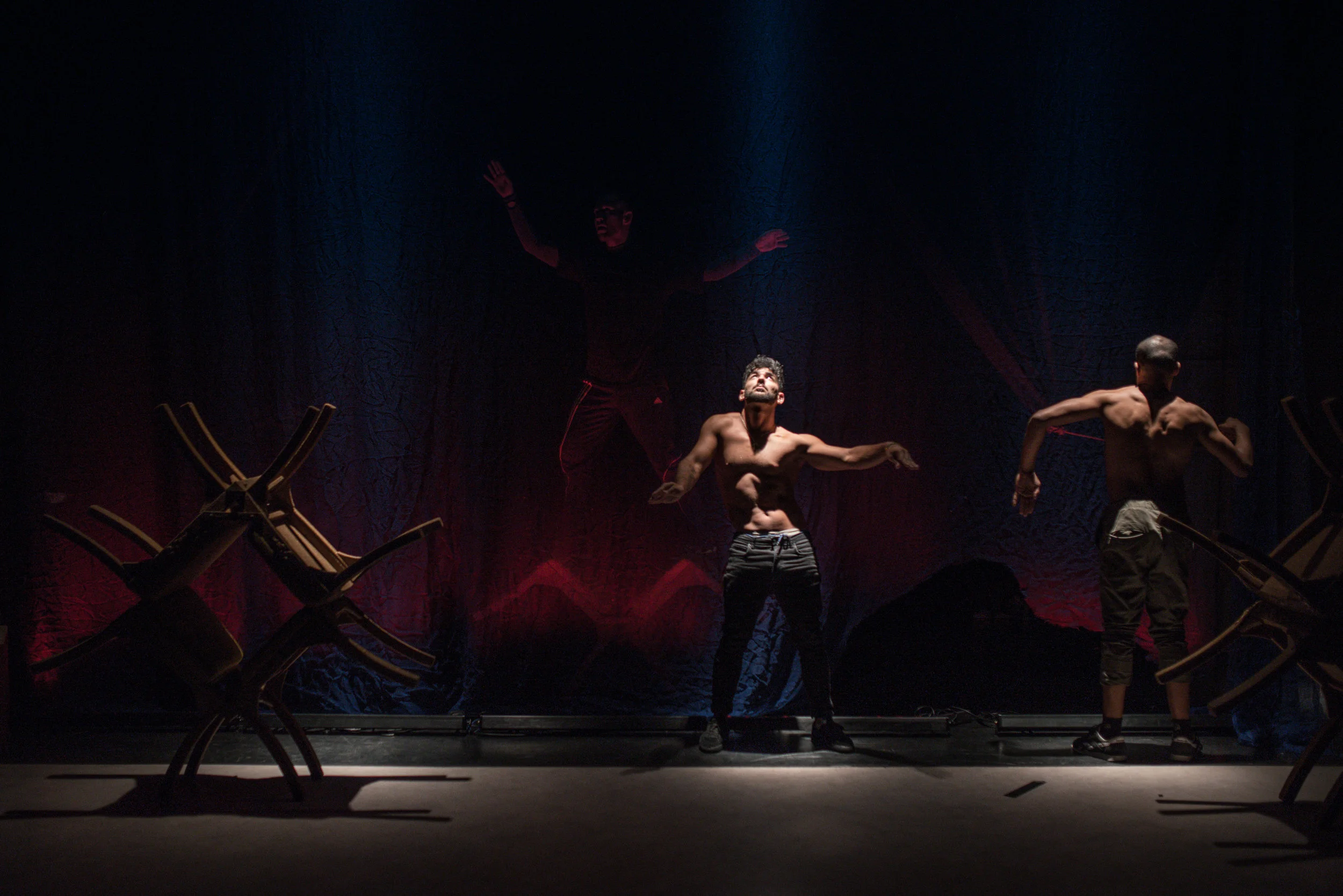 Trois danseurs nus en scène dans une lumière tamisée avec des chaises inversées.