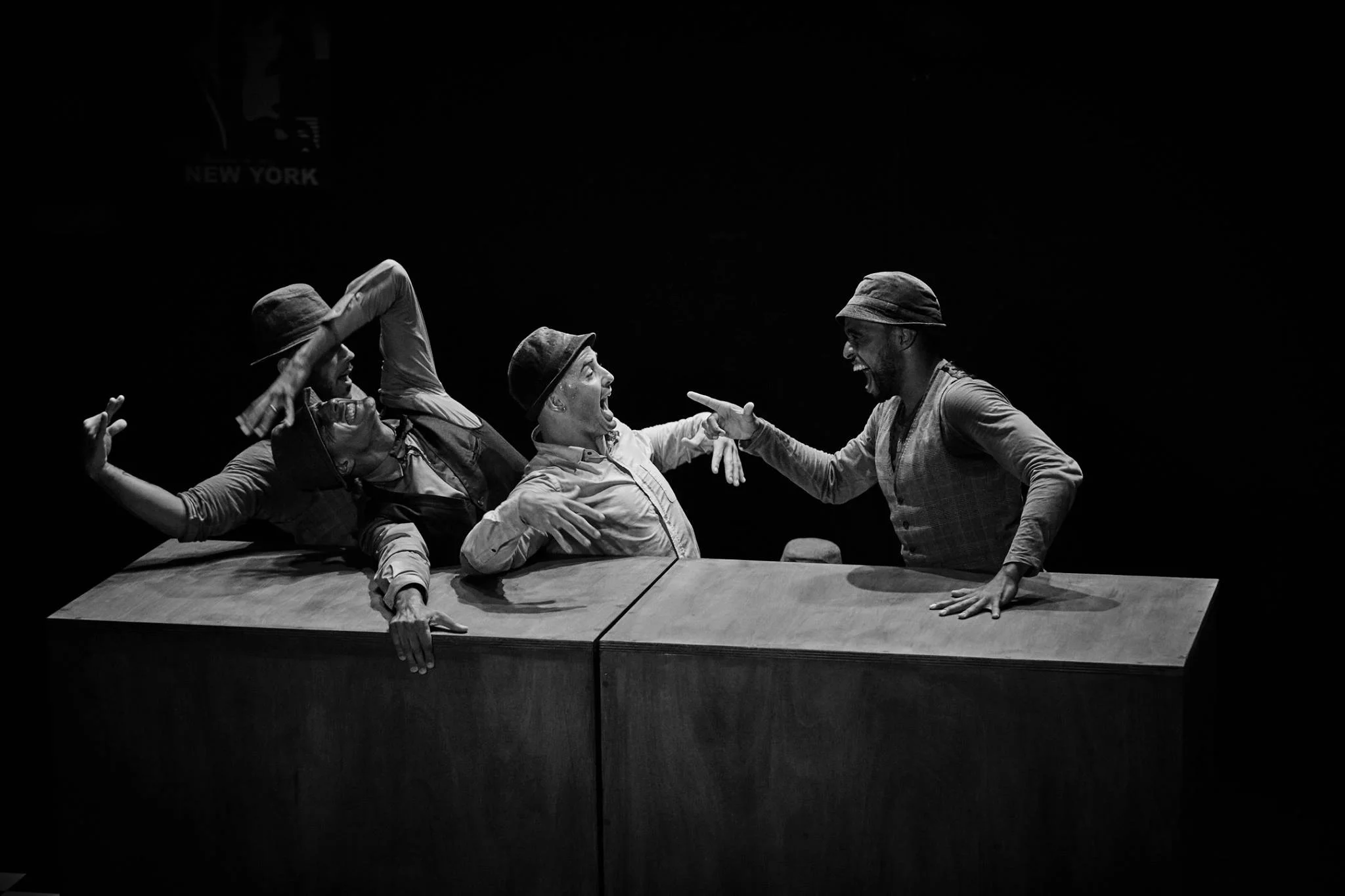 Quatre personnes faisant une scène théâtrale sur scène, deux tirant la langue et gesticulant, avec une table en premier plan, en noir et blanc