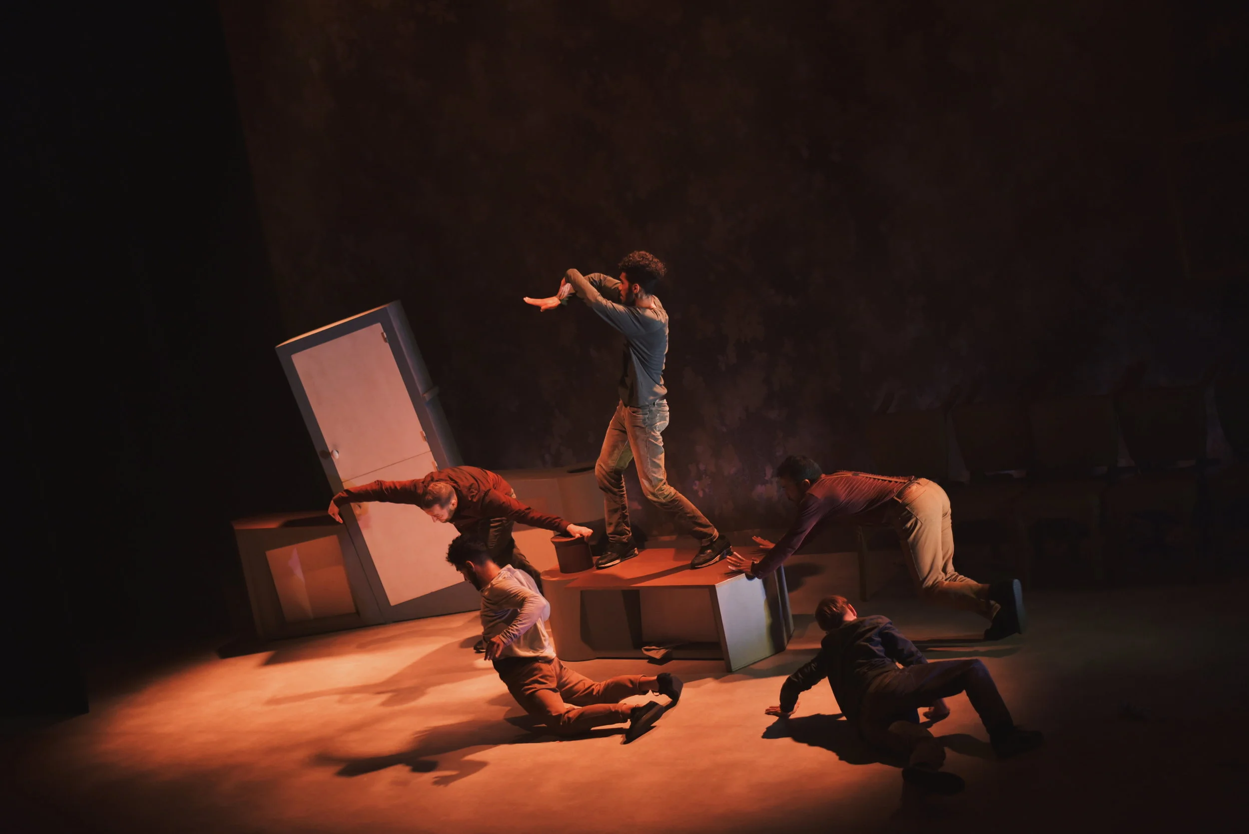 Un groupe d'acteurs en costume sur scène, en pleine représentation théâtrale ou danse moderne, avec un décor minimaliste comprenant des meubles.