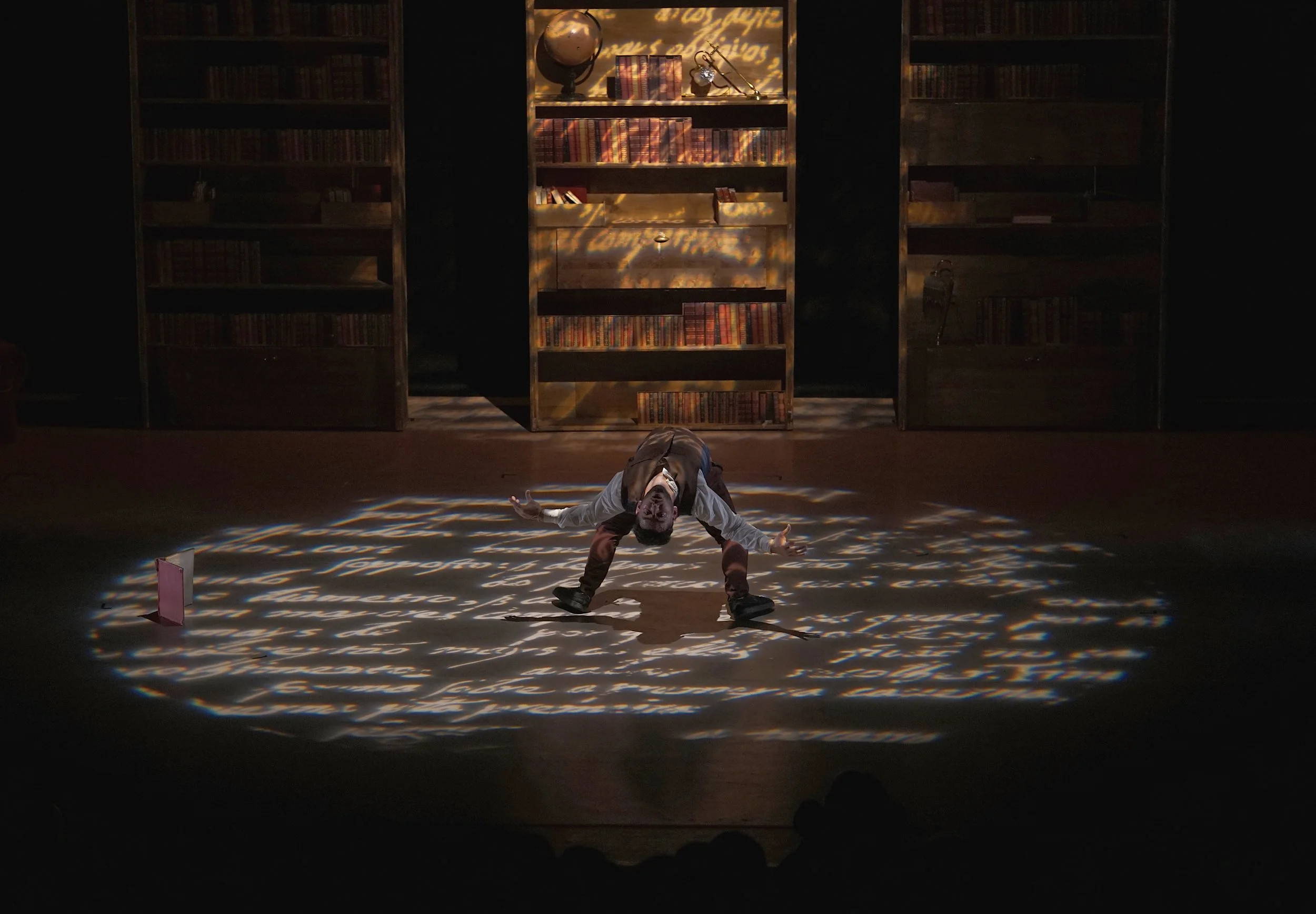 Une scène de théâtre avec un acteur au centre, entouré de livres, avec un éclairage de projection de texte sur le sol et derrière des étagères remplies de livres.