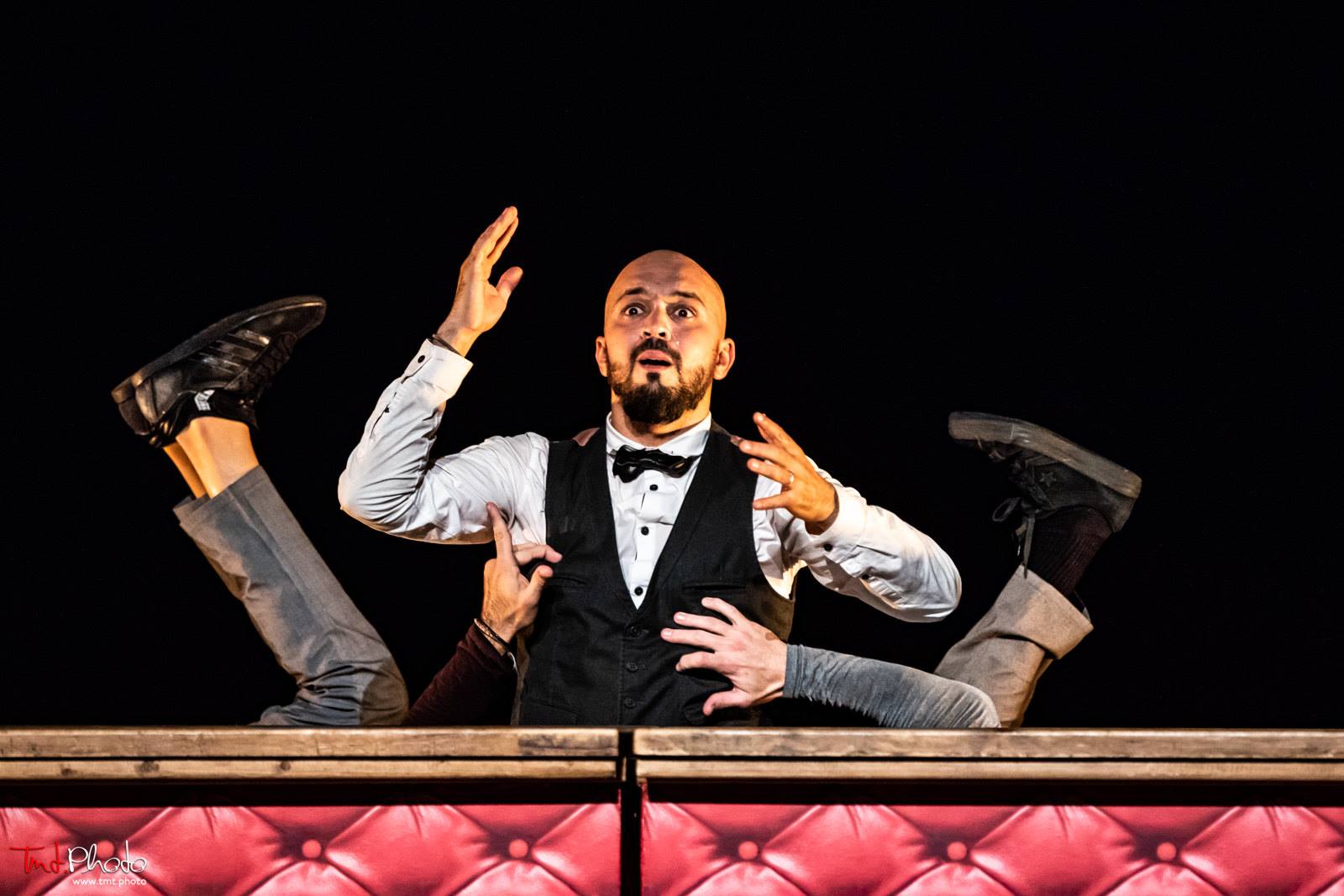 Une performance théâtrale avec un homme vêtu en veston noir et cravate, exprimant une émotion intense, entouré de plusieurs personnes dont seuls les bras sont visibles, dans un décor de scène en bois.