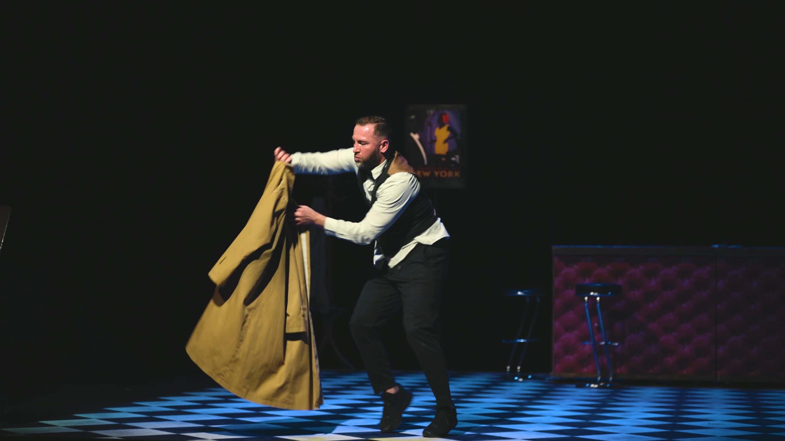 Un homme en scène, en train de retirer un manteau beige, dans un théâtre avec un sol en damier noir et blanc. Un tableau de New York en fond, et un meuble avec des fleurs rouges.