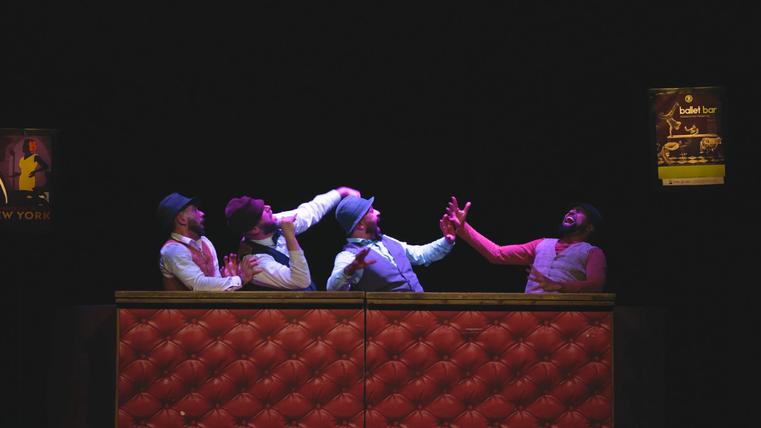 Cinq hommes en costumes vintage performants dans une pièce de théâtre, entre eux, ils semblent jouer une scène comique ou dramatique, avec une mise en scène sombre et des posters colorés en arrière-plan.