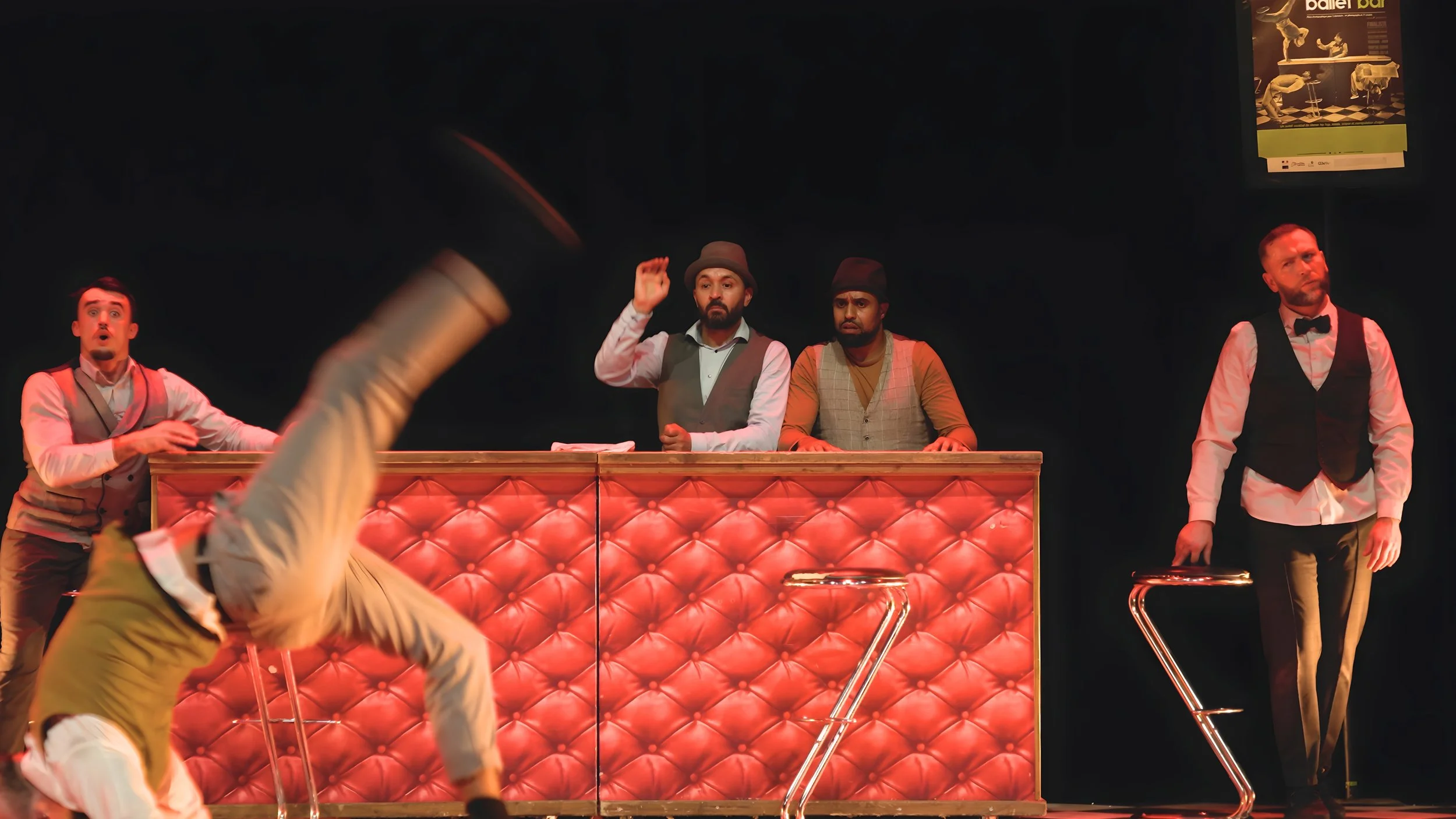 Quatre hommes en costumes de spectacle, deux derrière un bar rose rembourré, un à gauche effectuant une acrobatie, et un à droite en train de se tenir debout, lors d'une performance théâtrale ou un spectacle musical.