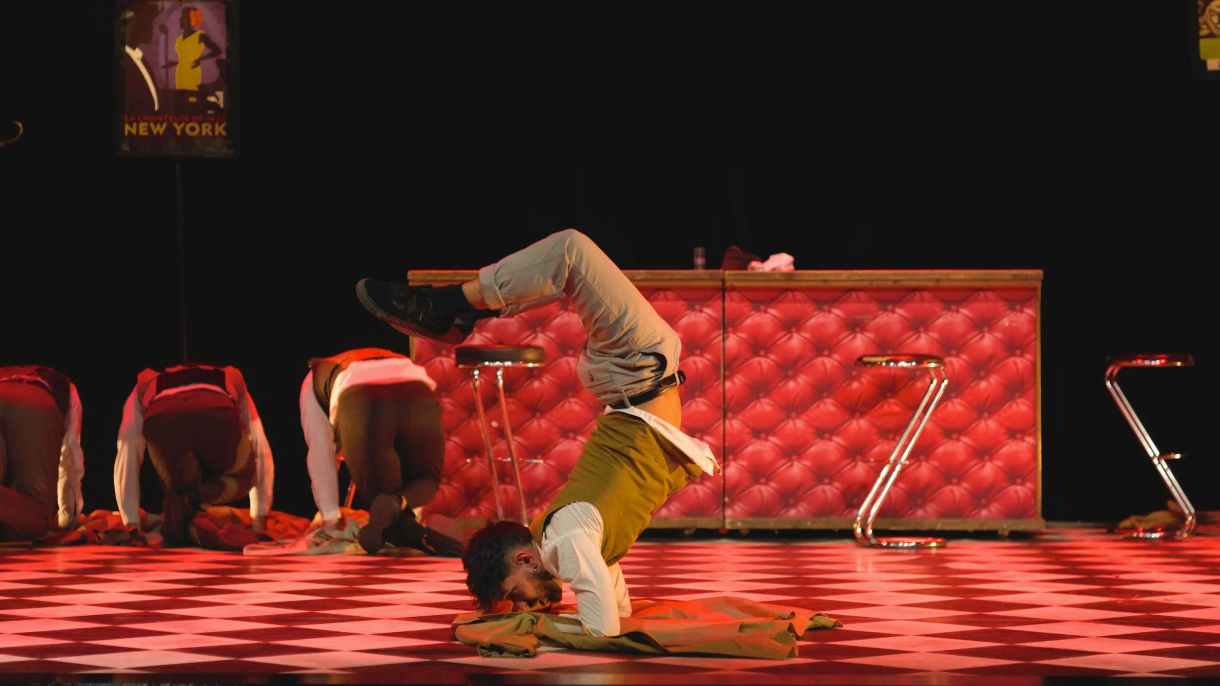 Un homme en équilibre sur la tête, faisant un headstand dans une scène de théâtre avec un sol en damier noir et blanc, un canapé rouge en arrière-plan, et plusieurs autres personnes à genoux ou pliées.