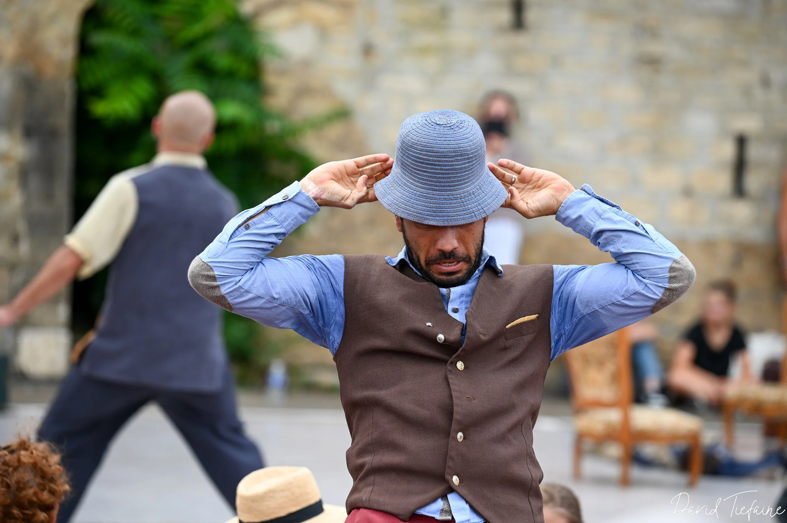 Un homme portant un chapeau bleu et un gilet marron ajuste son chapeau, avec d'autres personnes en arrière-plan dans une scène extérieure. 