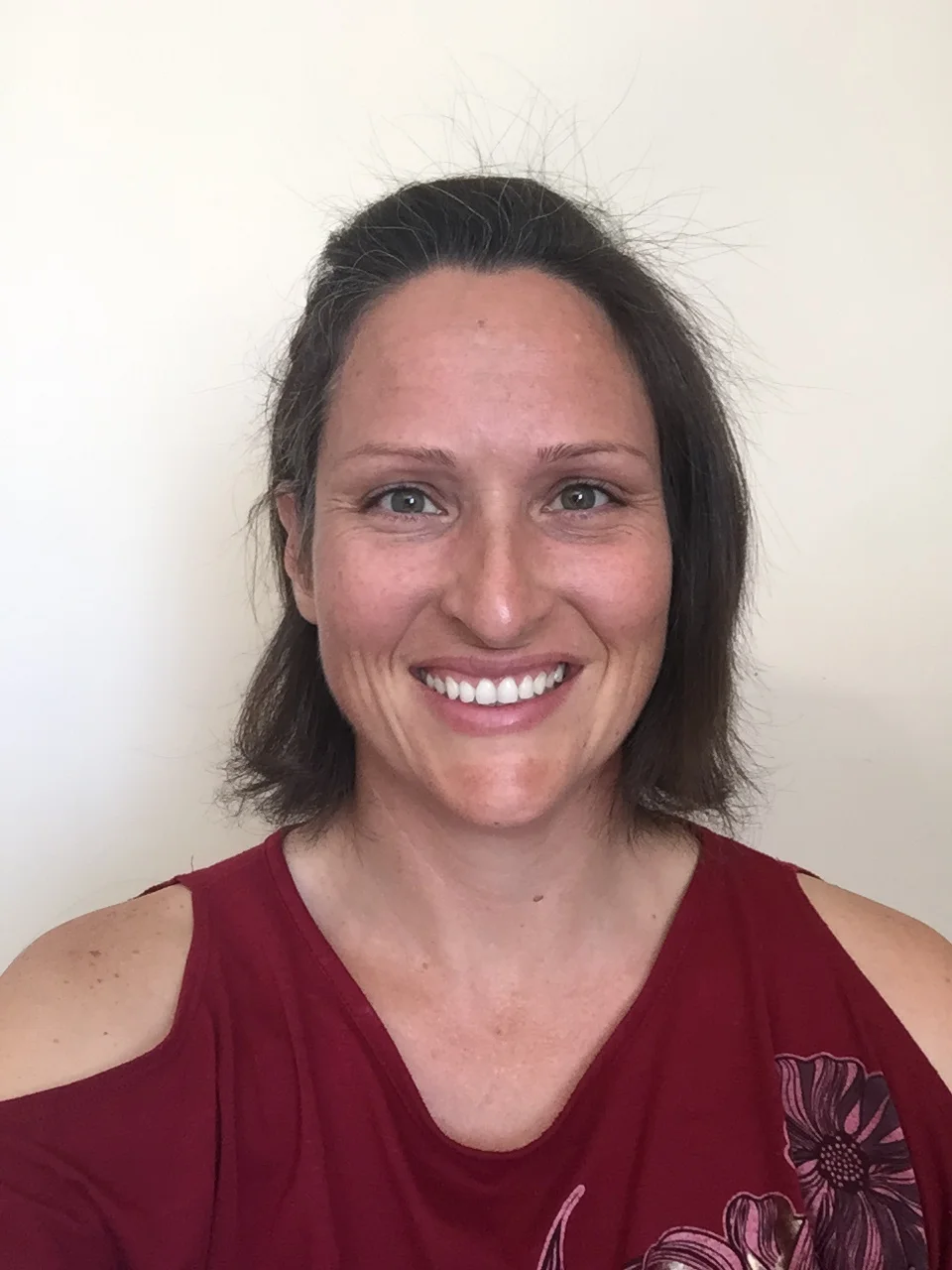 Close-up of a woman with shoulder-length brown hair, wearing a red top with cold-shoulder cutouts and a floral design, smiling against a plain white wall.