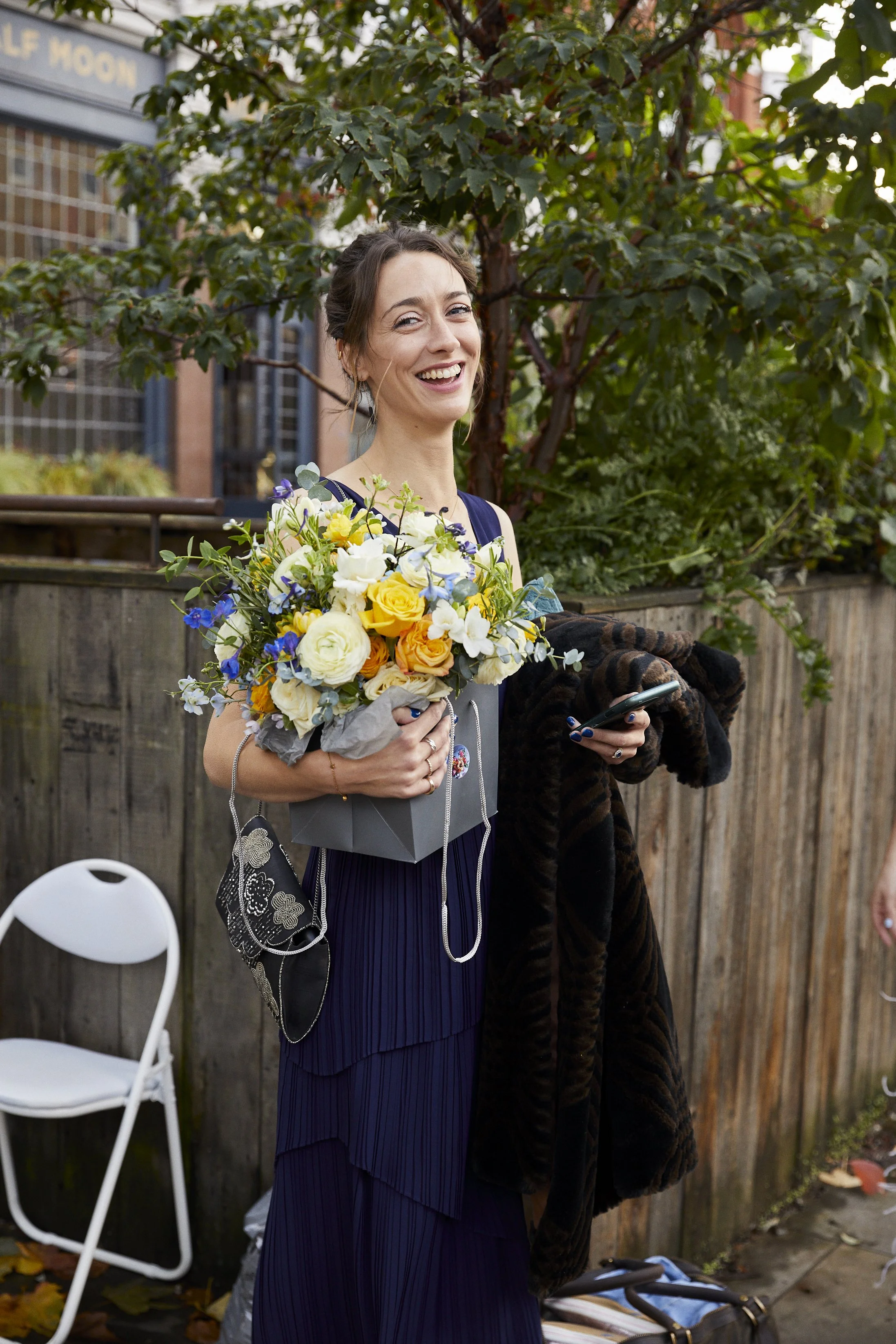 A woman smiling and holding a colorful bouquet of flowers outside next to a wooden fence and trees, wearing a dark blue dress and holding a cellphone, with a white chair nearby.