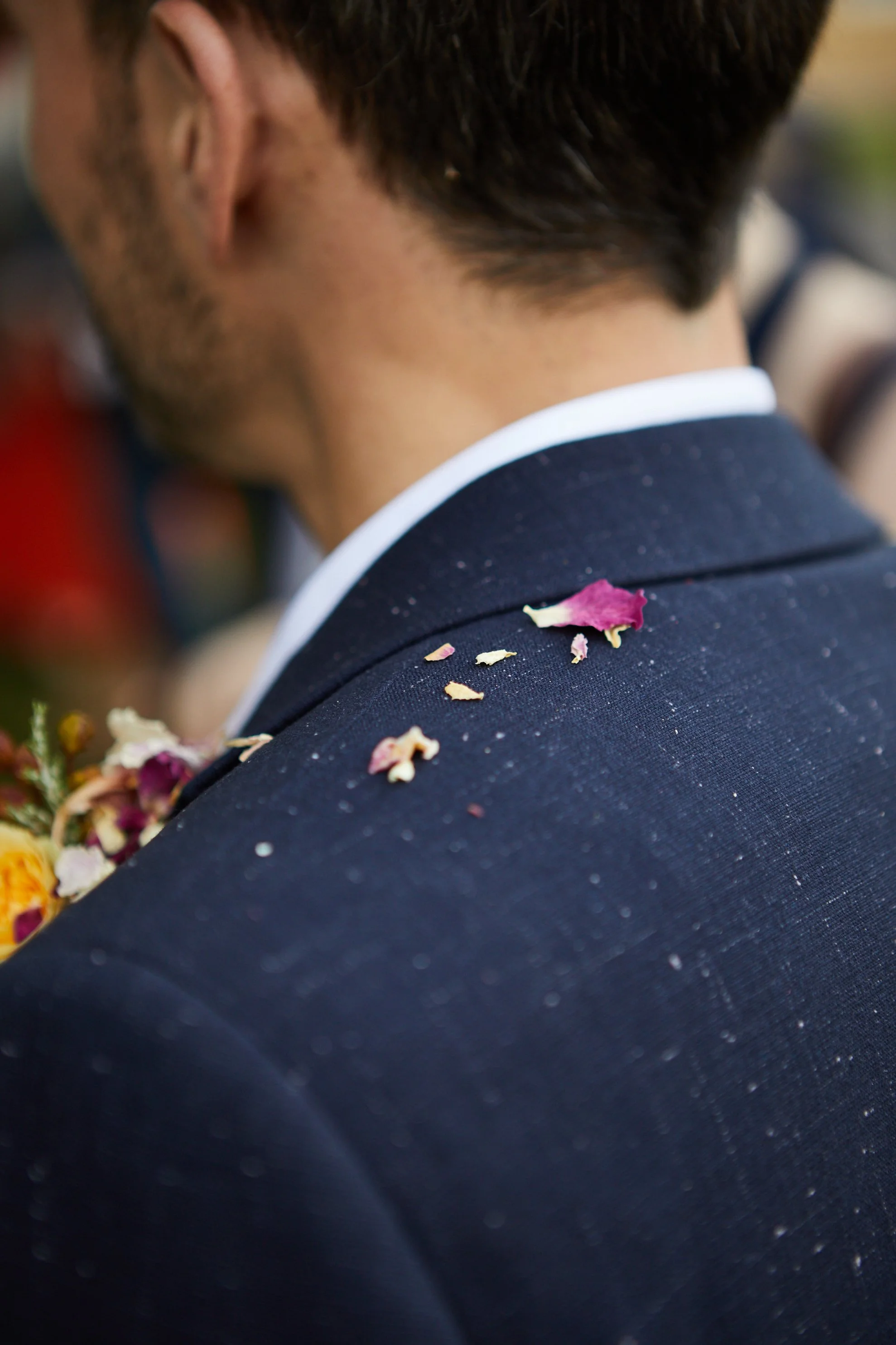 Close-up of a man's shoulder and back, showing a dark suit jacket with flower petals on it, at what appears to be a wedding or formal event.