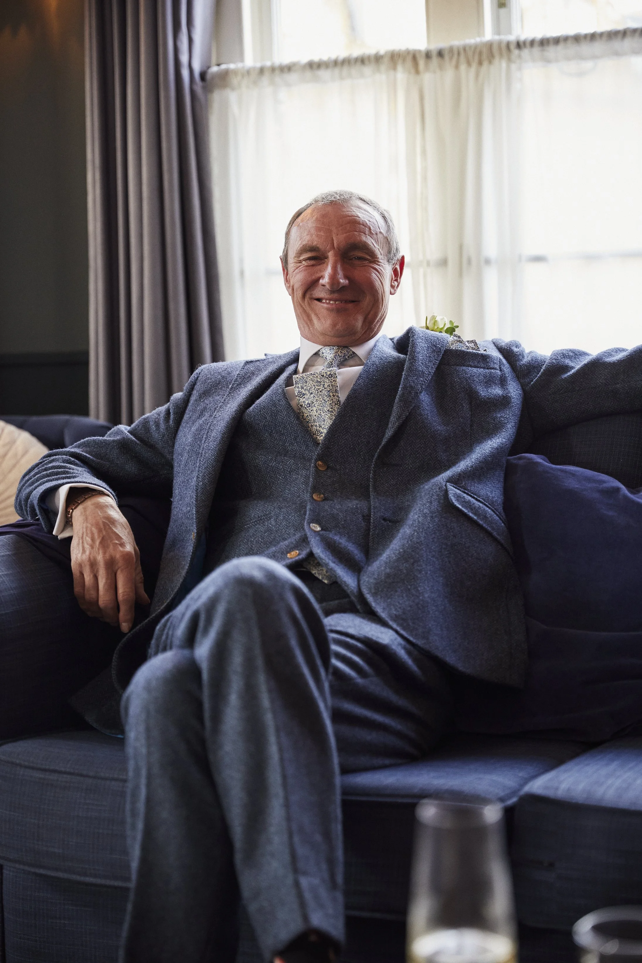 A smiling man in a gray suit and tie sitting on a dark-colored sofa in a well-lit room with large windows and curtains.