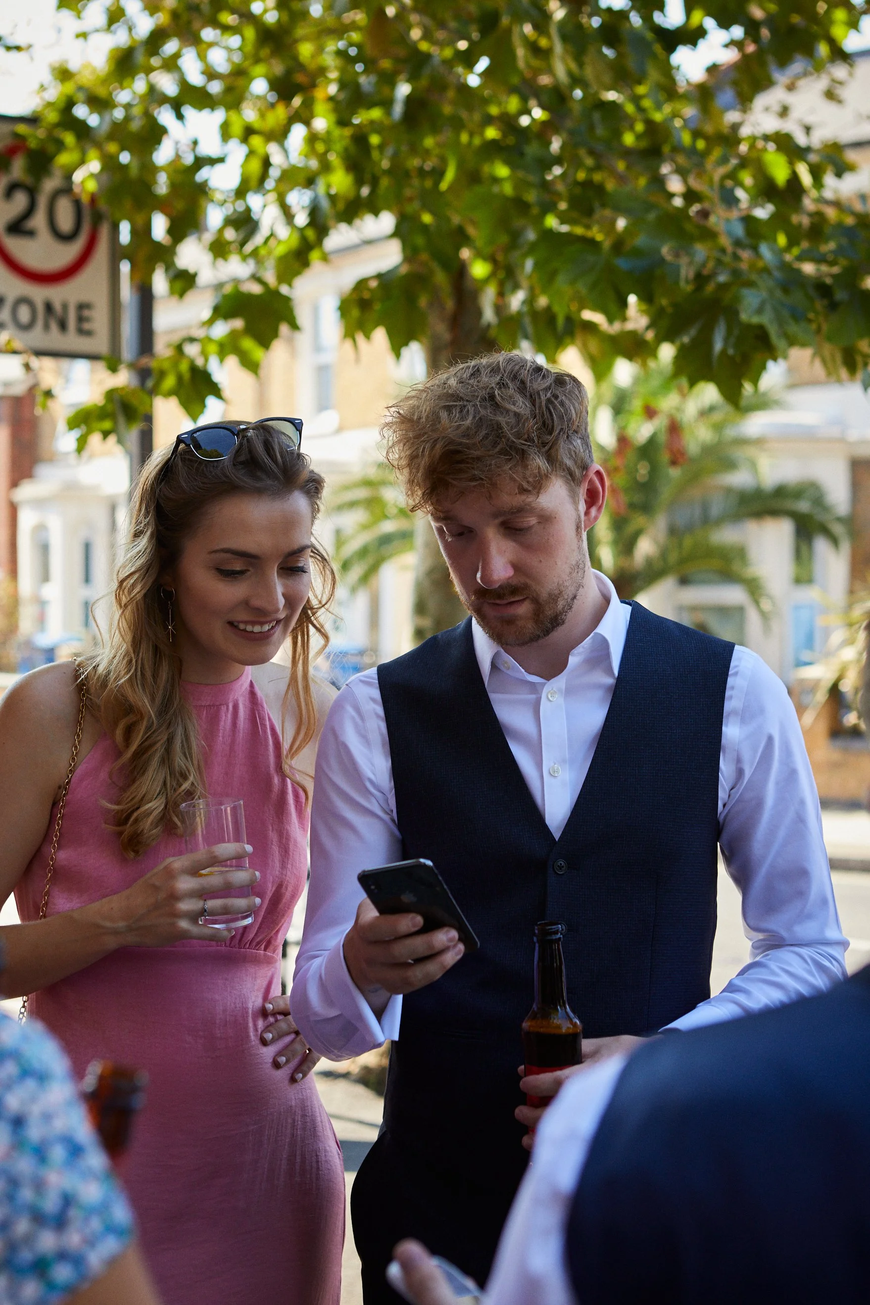 A man in a white shirt and black vest looking at his phone while holding a beer bottle, a woman in a pink dress standing beside him, holding a glass of drink, both outdoors on a sunny day with trees and buildings in the background.