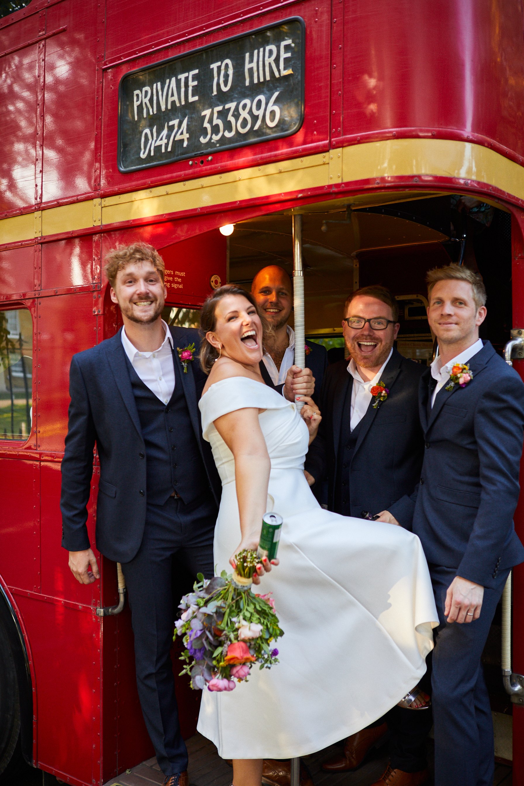 Group of five people in wedding attire, celebrating in front of a vintage red double-decker bus, with a sign that reads 'Private to Hire' and a phone number.