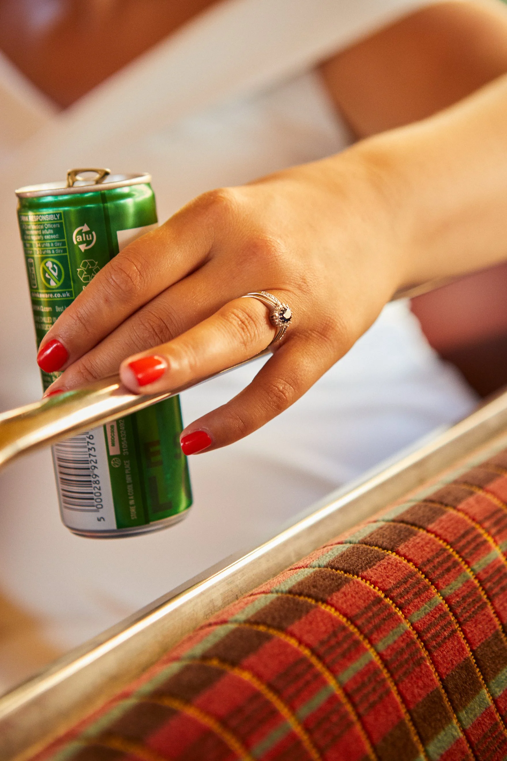 Close-up of a woman's hand with red nails holding a canned drink, wearing a ring, and resting on a metallic surface near a colorful, checked upholstery.