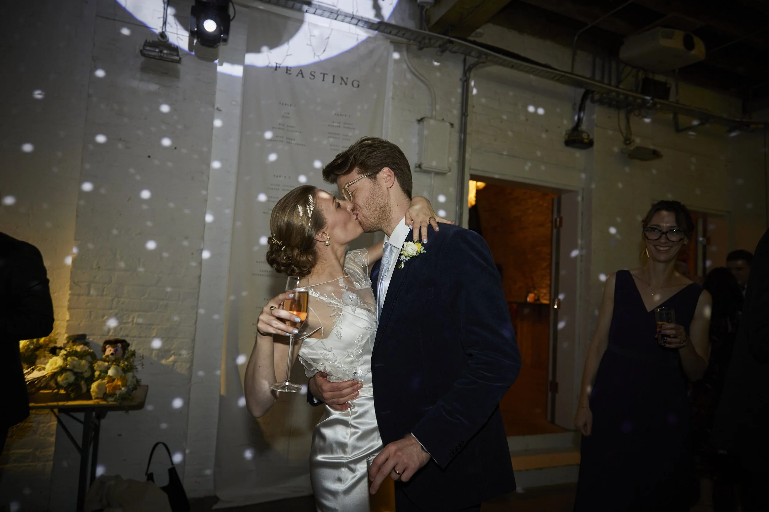 A bride and groom share a kiss at their wedding reception, with the bride holding a glass of champagne and guests in the background celebrating.