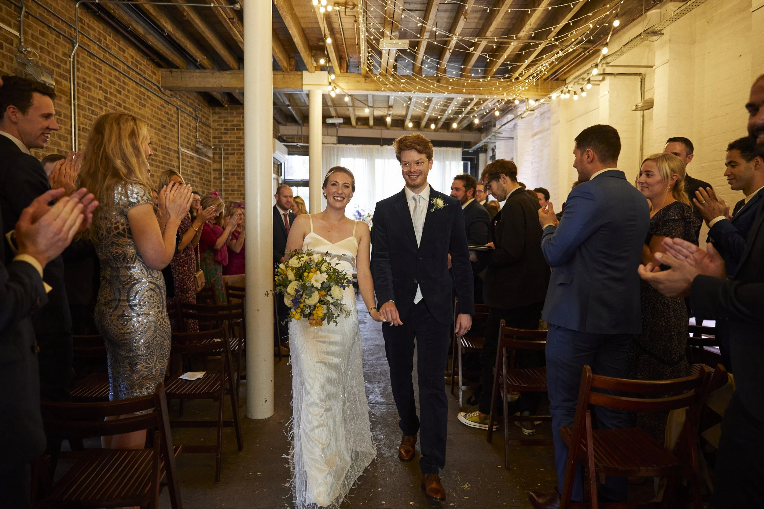 A newlywed couple walks down the aisle inside an industrial-style venue, smiling and holding hands, surrounded by clapping guests. The bride wears a white dress and holds a bouquet of flowers, while the groom wears a dark suit and glasses. Guests are