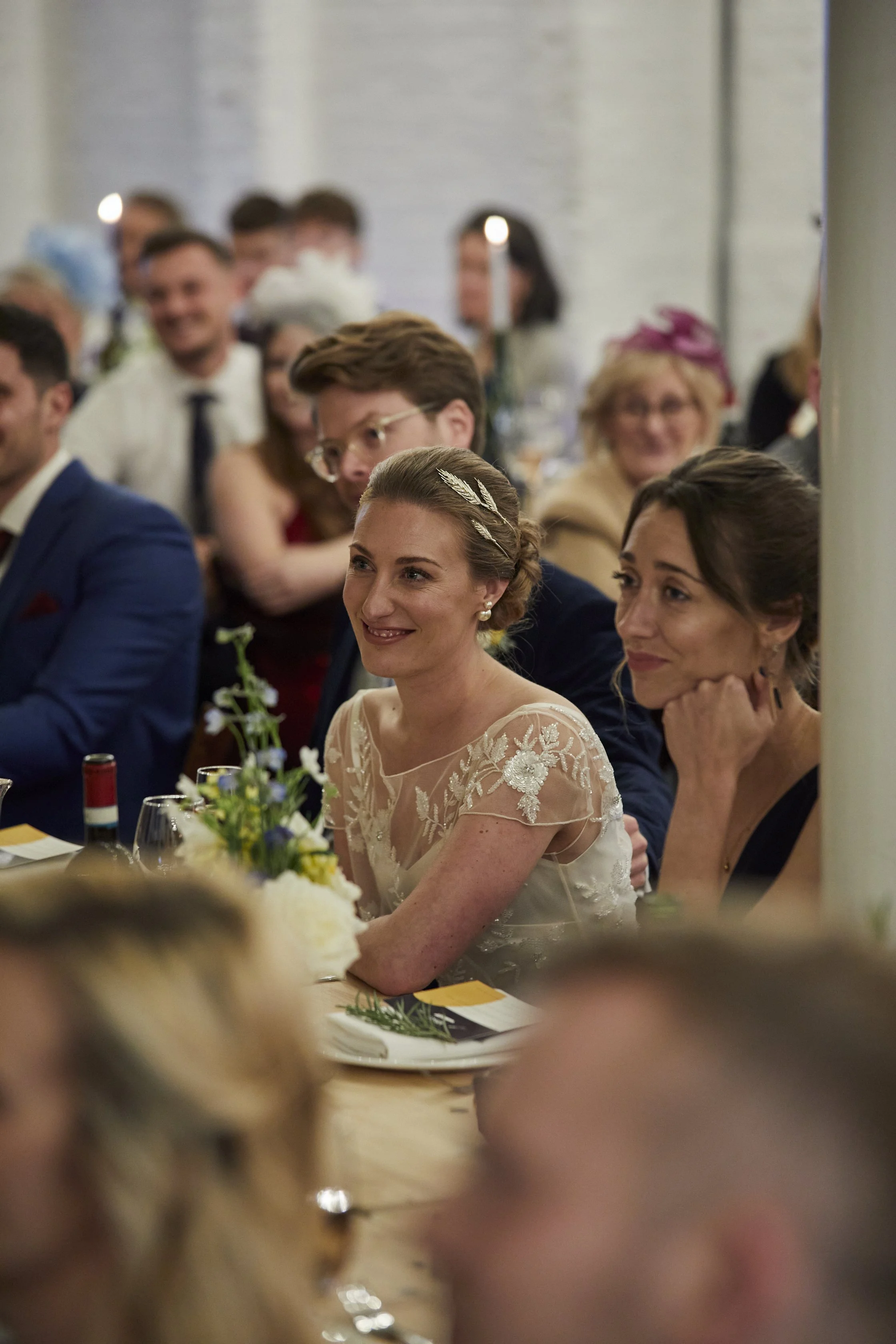 A woman in a cream lace wedding dress with floral embroidery and pearl earrings, smiling and sitting at a table during her wedding reception, surrounded by guests in formal attire.
