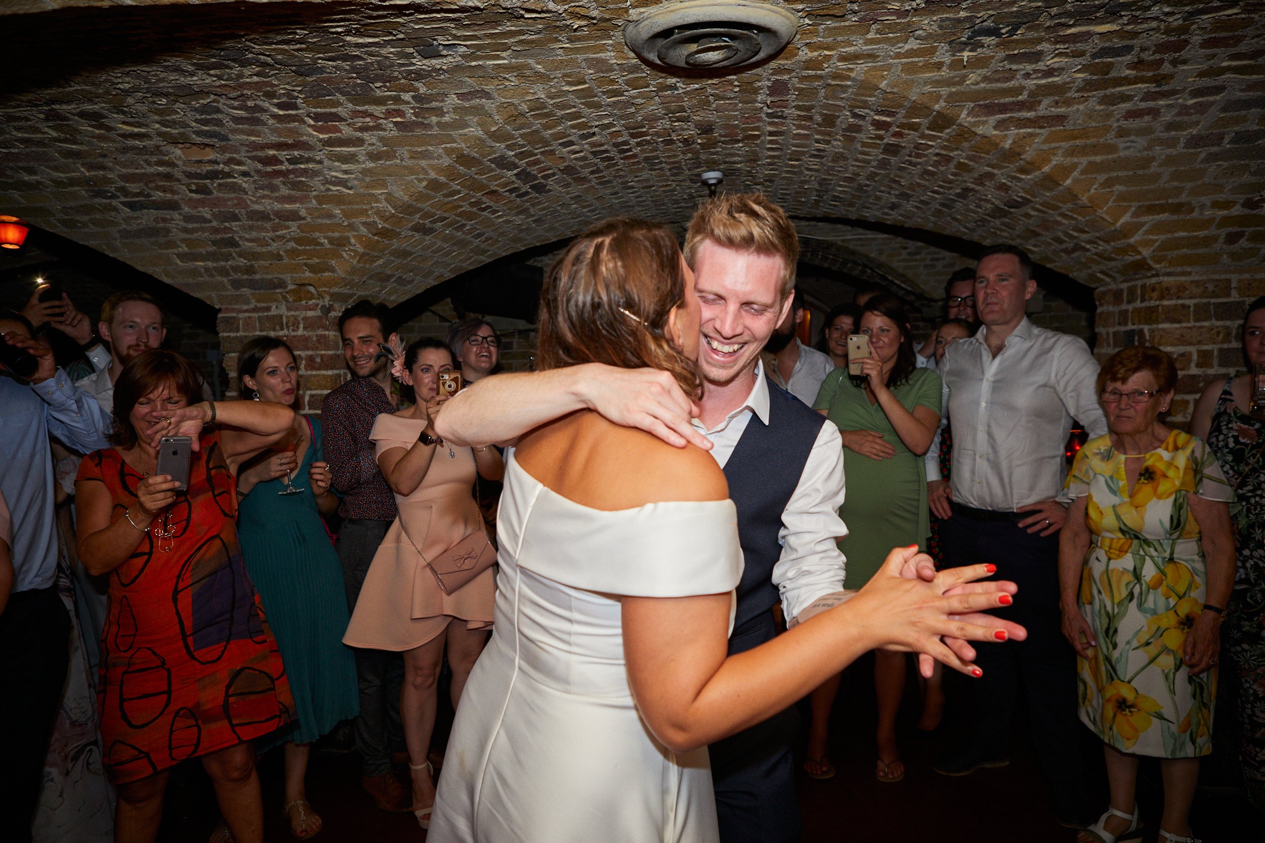 A couple dancing closely, with the woman wearing an off-the-shoulder white dress and the man in a white shirt and dark vest, surrounded by smiling guests in colorful attire, in a brick-vaulted venue.