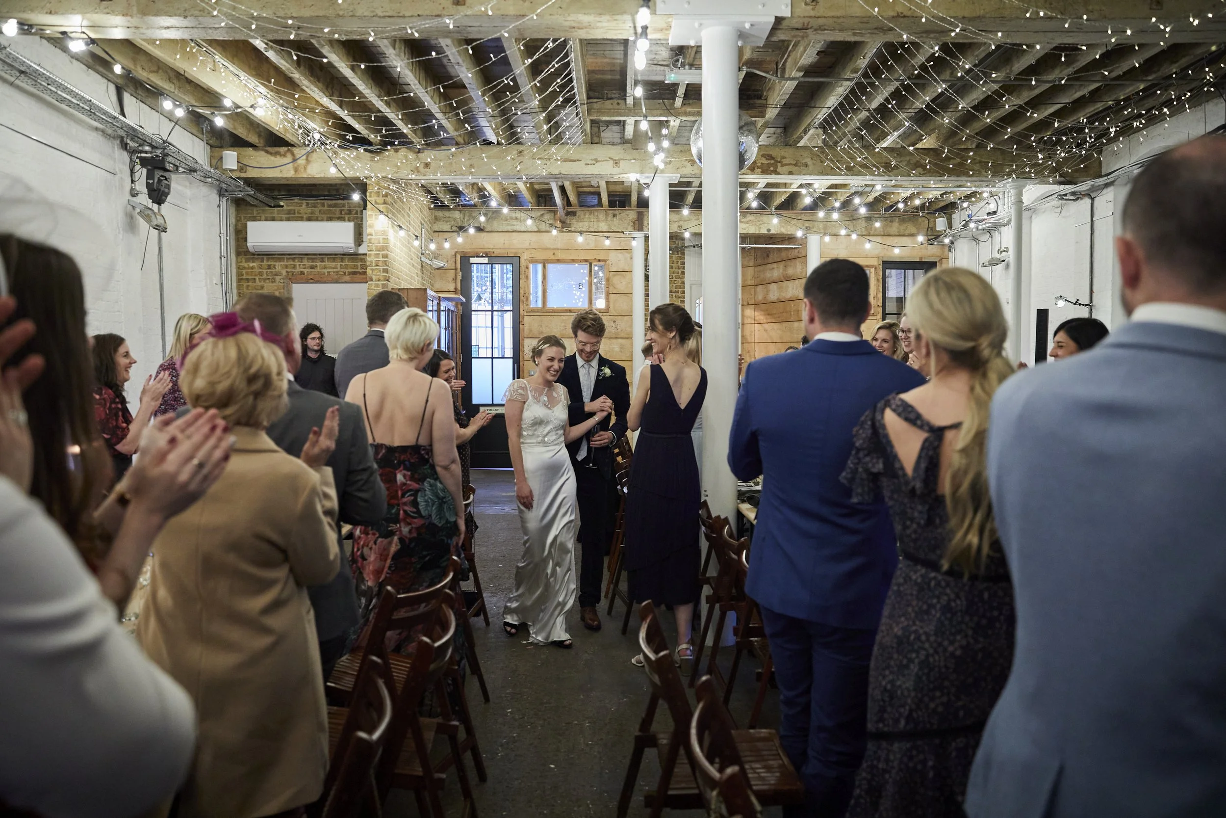 Wedding reception with guests standing and clapping, a bride and groom walking down the aisle with other guests seated and standing around, decorated with string lights and rustic decor.
