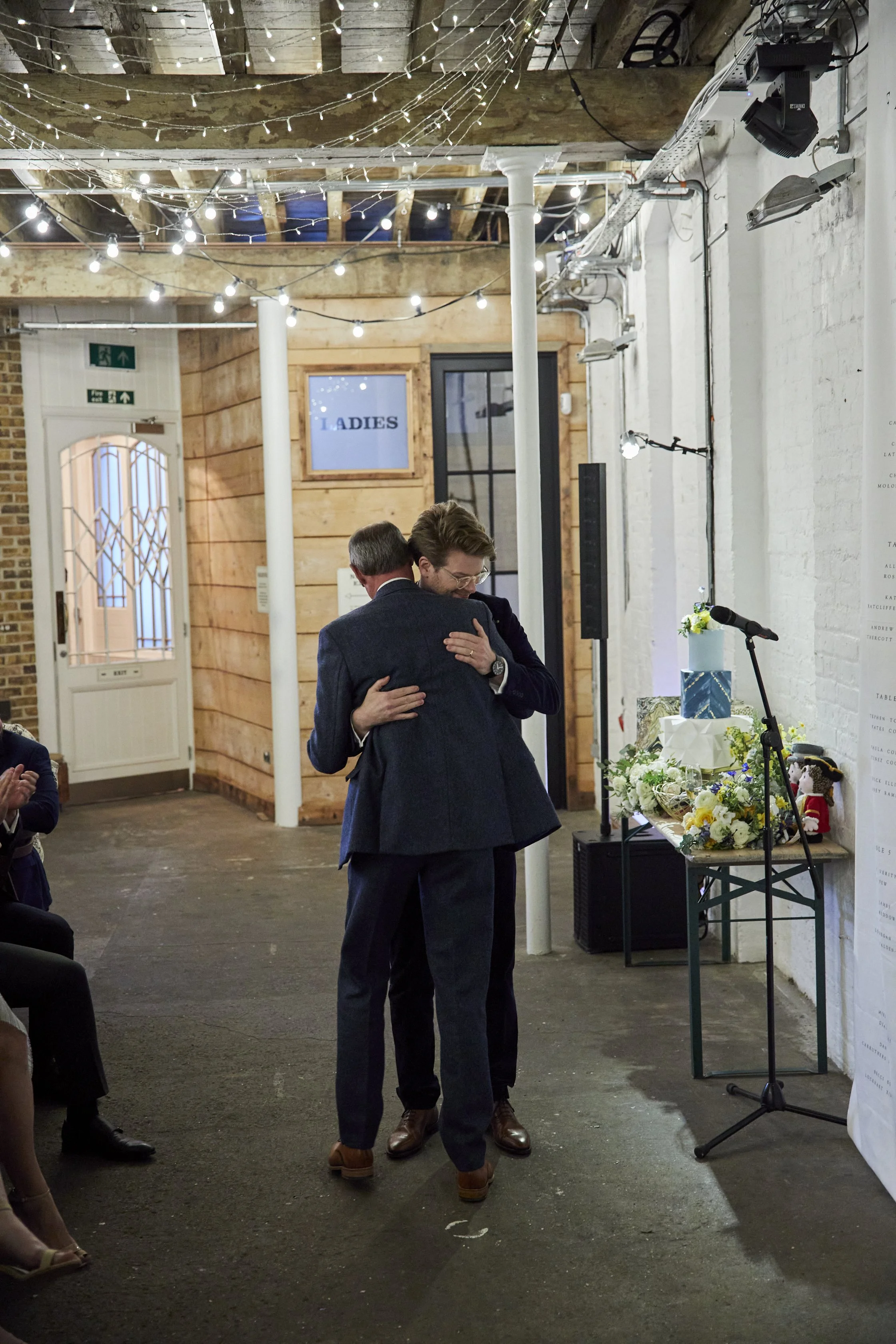 Two men in suits hugging in a rustic indoor setting with string lights, a microphone, and floral arrangements, likely during a celebration or ceremony.