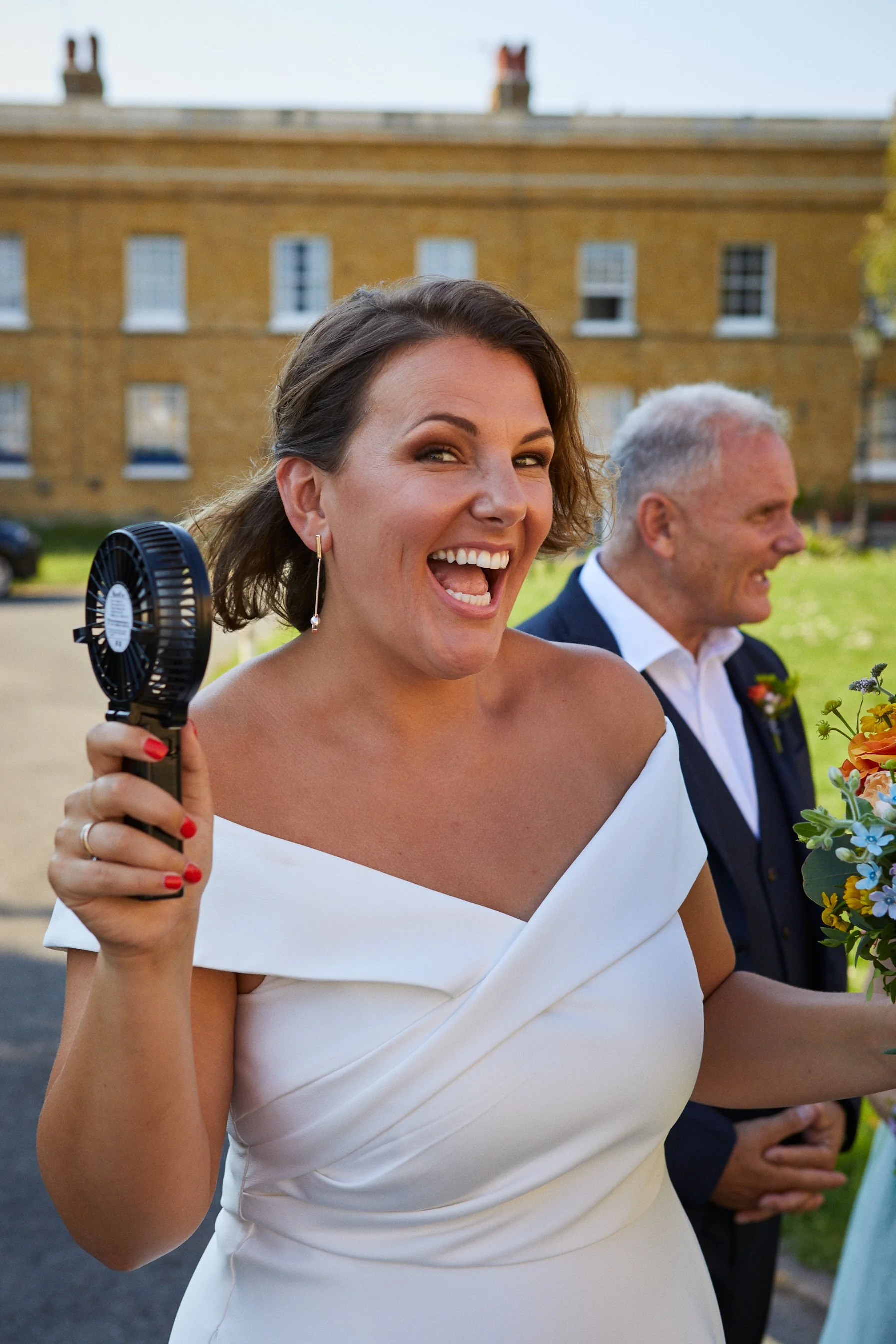 Woman in white dress holding a small fan, smiling and appearing happy at an outdoor event, likely a wedding, with a man in a tuxedo and a flower arrangement visible.