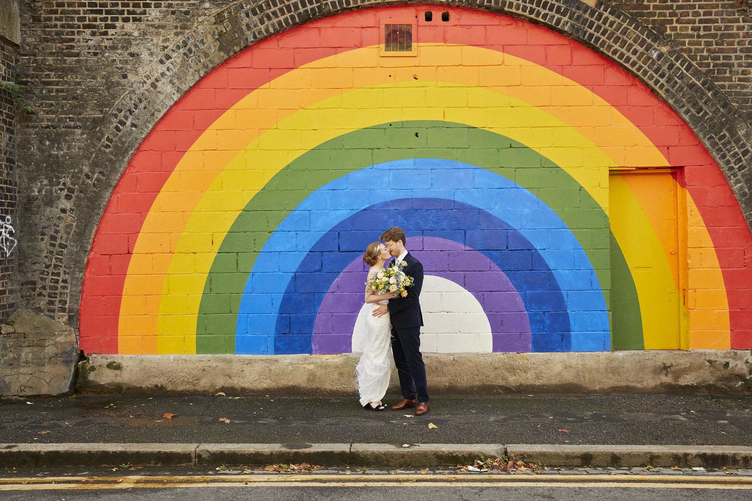 A couple in wedding attire standing close and sharing a kiss in front of a colorful rainbow mural painted on a brick wall.