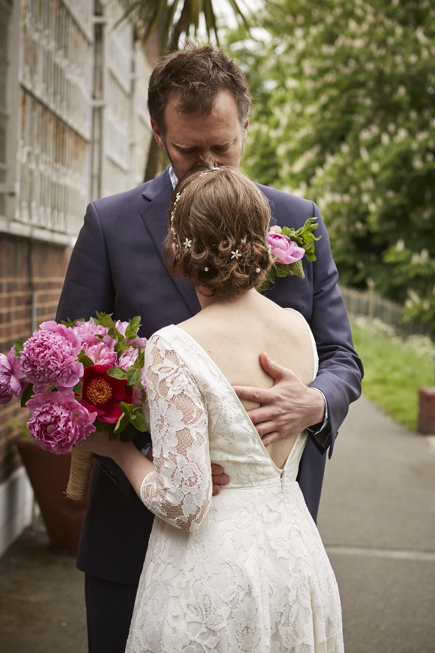A groom in a suit holding a bride in a lace wedding dress, with both holding pink flowers, sharing an intimate moment outdoors.