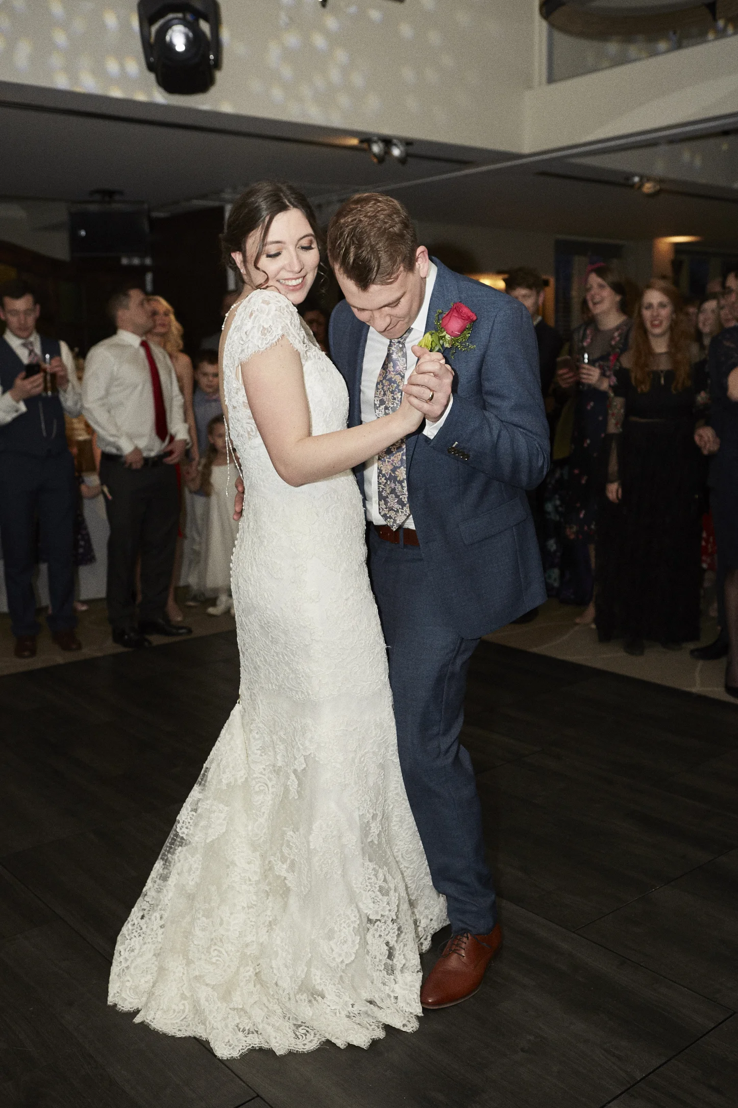 A bride and groom dance together at their wedding reception while guests watch and smile in the background.