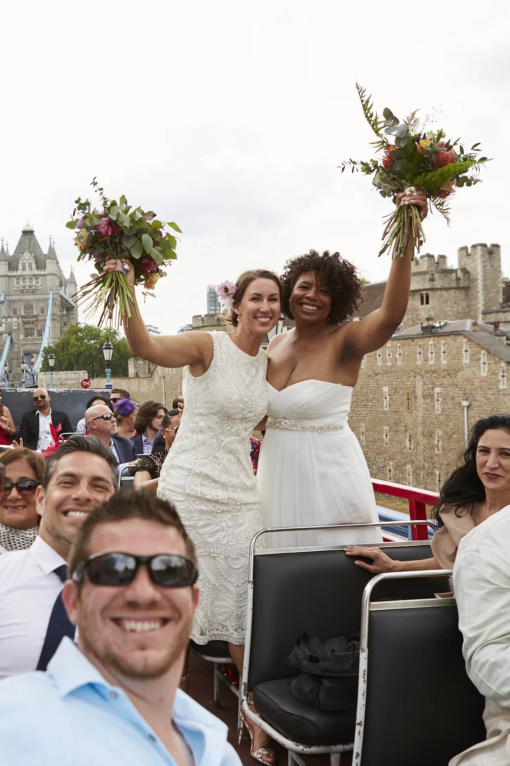 Two women, one with light skin and one with dark skin, standing on a boat, celebrating a wedding with bouquets of flowers raised in the air, with a historic castle and cityscape in the background, along with several smiling guests.