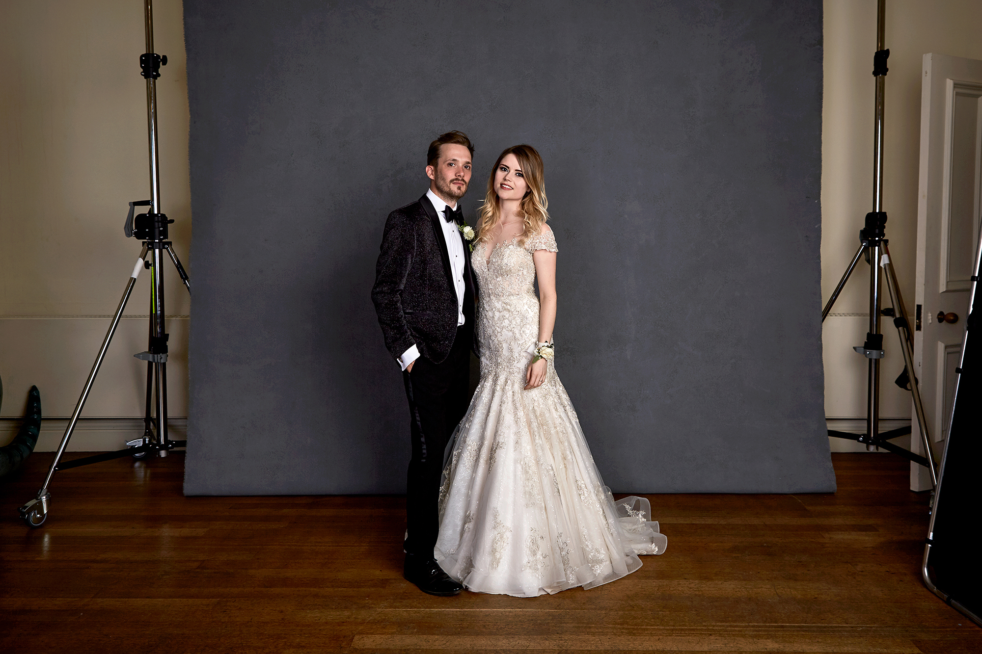 A bride and groom in formal wedding attire standing together in front of a gray backdrop during a photoshoot.