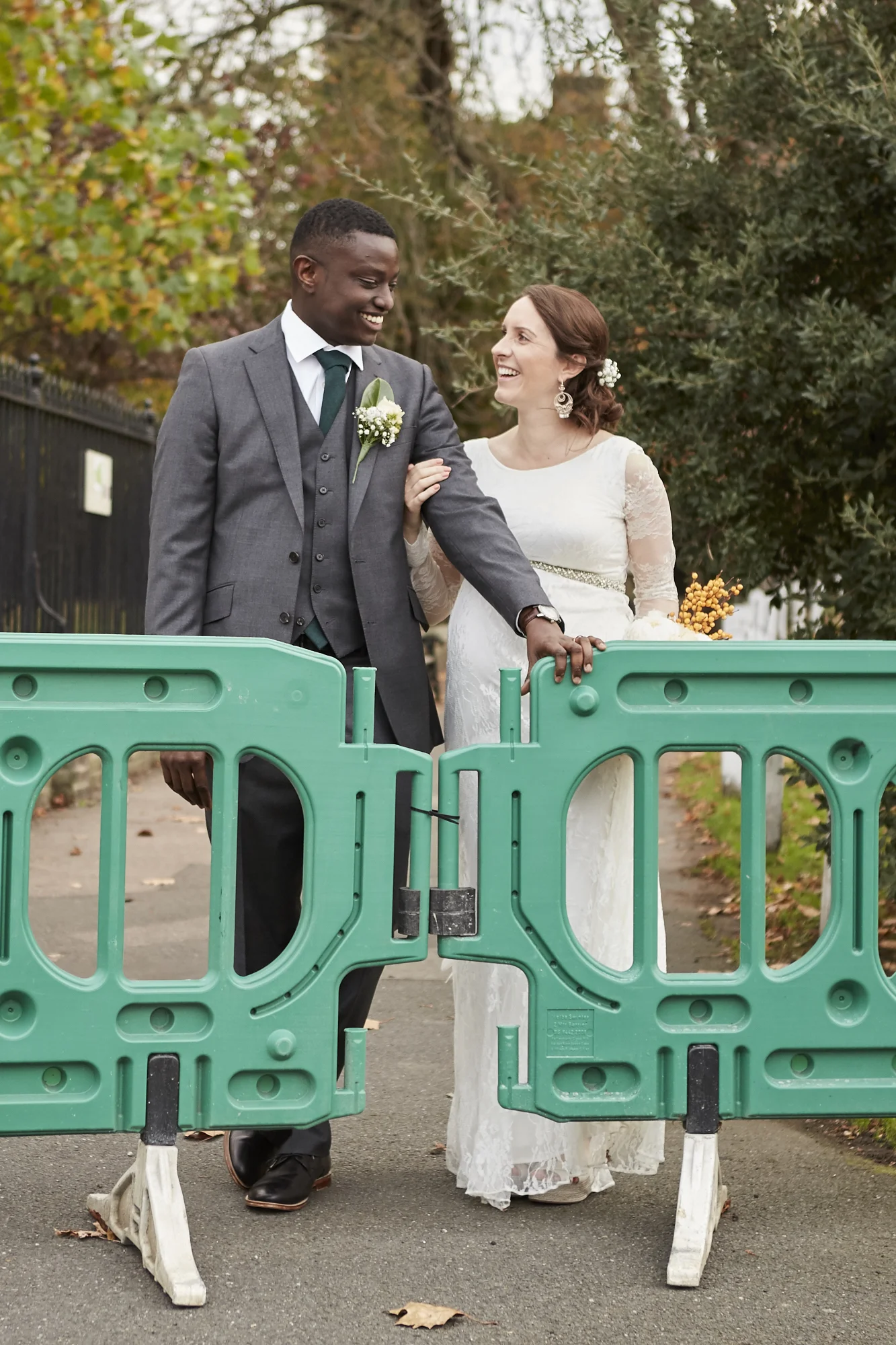 A bride and groom in wedding attire holding hands behind green safety barriers outdoors, with trees and fall foliage in the background.