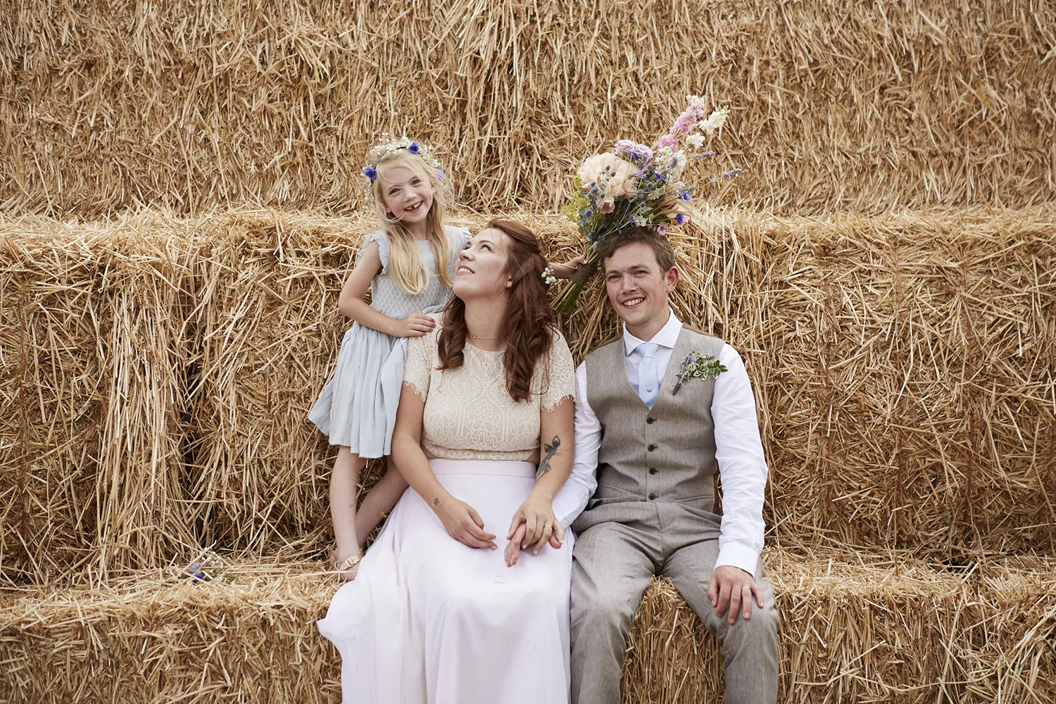 A family celebrating a wedding in front of a straw bale backdrop, with a woman and a man sitting and a young girl standing beside them, holding a bouquet of flowers.