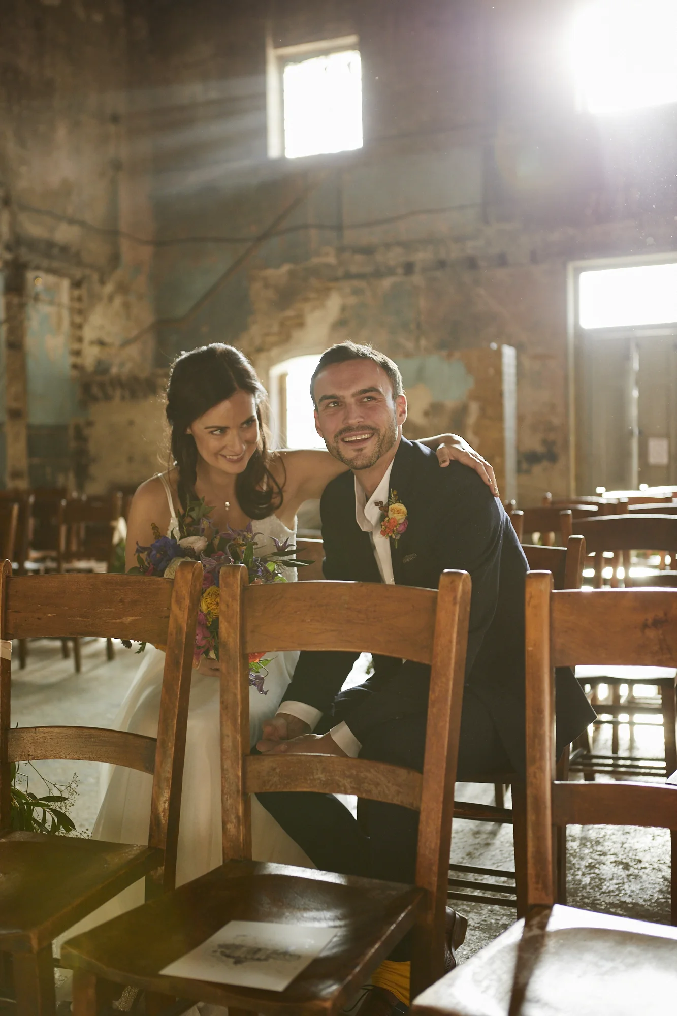 A couple posing for a wedding photo inside a rustic, old brick building with sunlight streaming through the windows. The woman is wearing a white wedding dress, and the man is dressed in a dark suit. They are smiling and leaning close to each other, with the woman holding a bouquet of flowers.