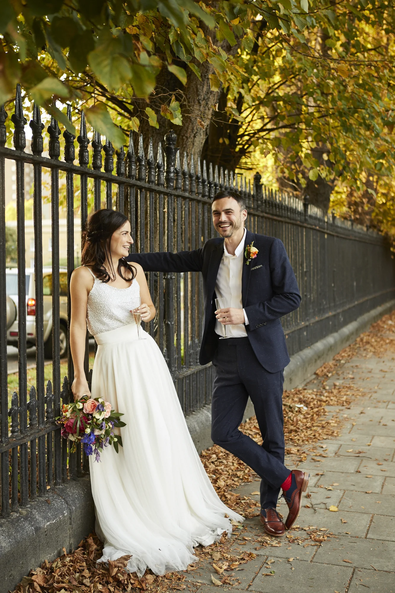 A newlywed couple standing by a black iron fence on a sidewalk covered with autumn leaves, smiling and having a conversation in the late afternoon or early evening.