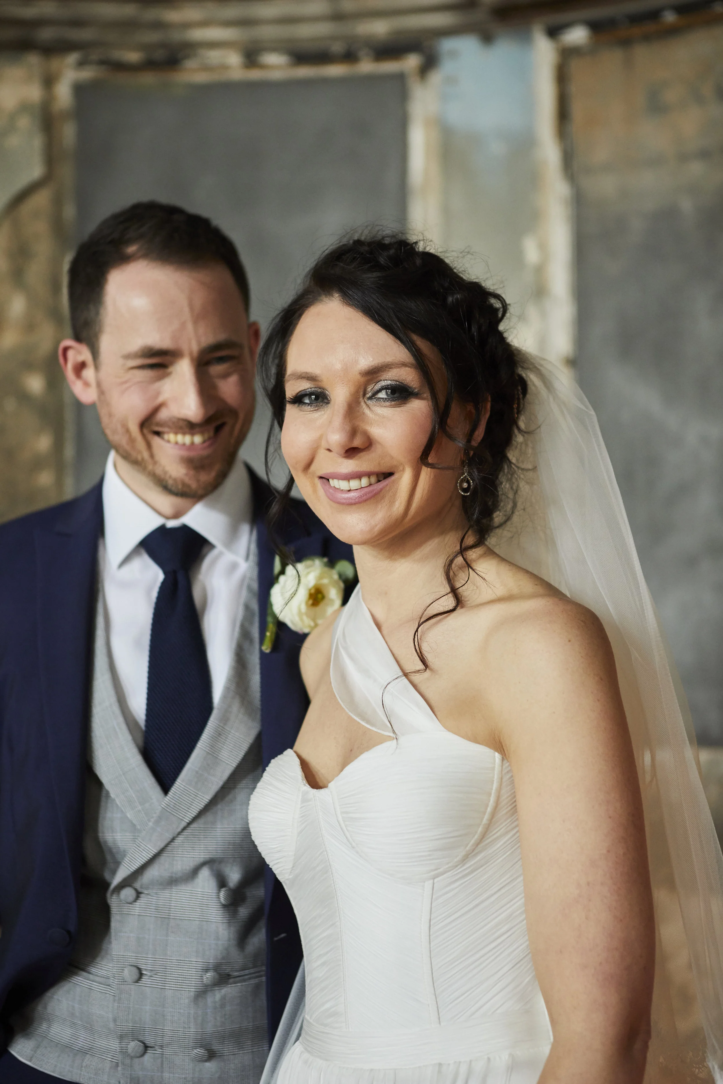 Smiling bride in a white wedding dress with dark hair and a veil, and smiling groom in a dark suit with a blue tie, standing together in an indoor setting.