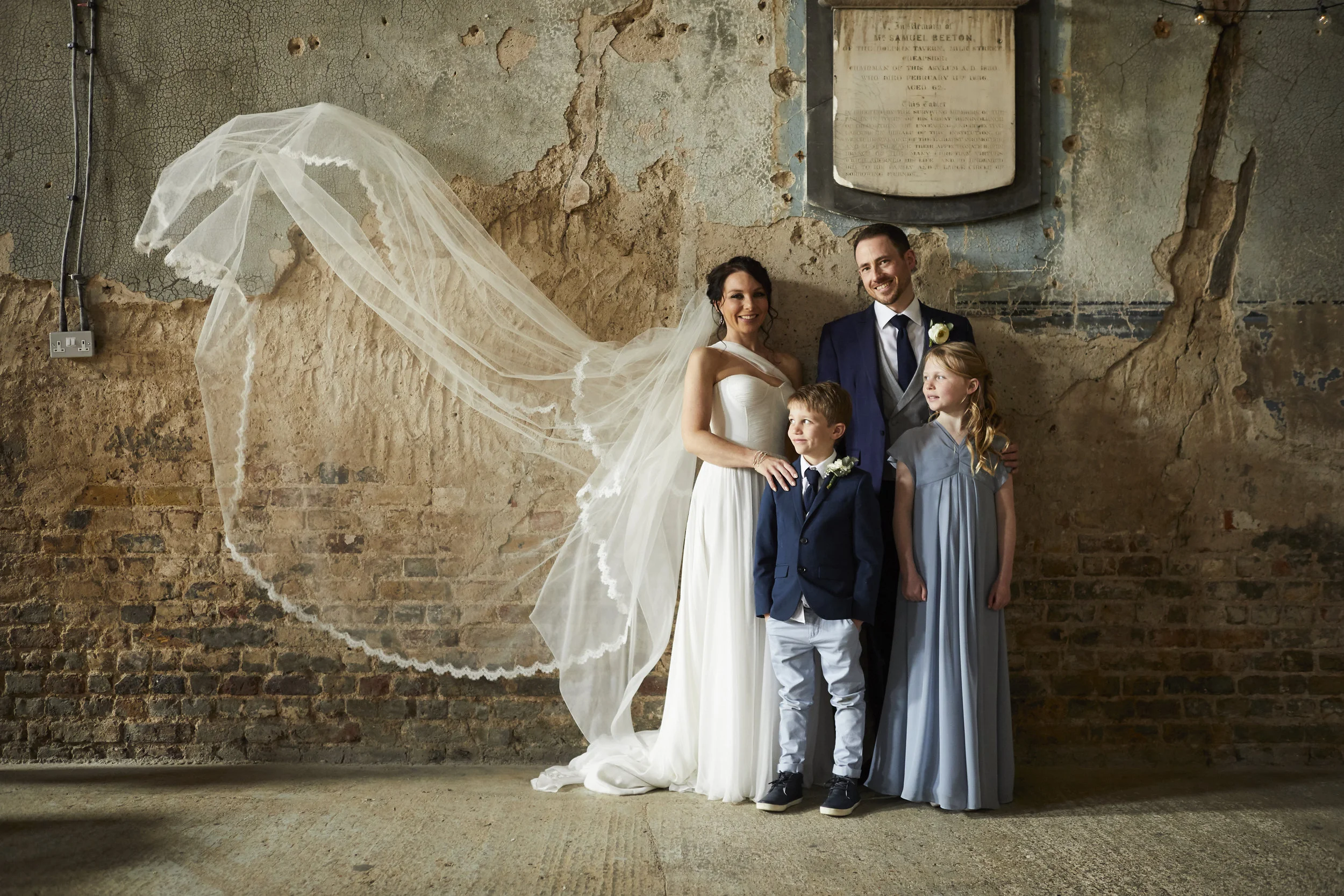 Wedding couple with two children standing against a weathered brick and plaster wall. Bride in white gown with veil, groom in navy suit with boutonniere. Young boy and girl dressed in formal attire. The wall behind them has peeling paint and exposed brick, with a framed memorial plaque above.