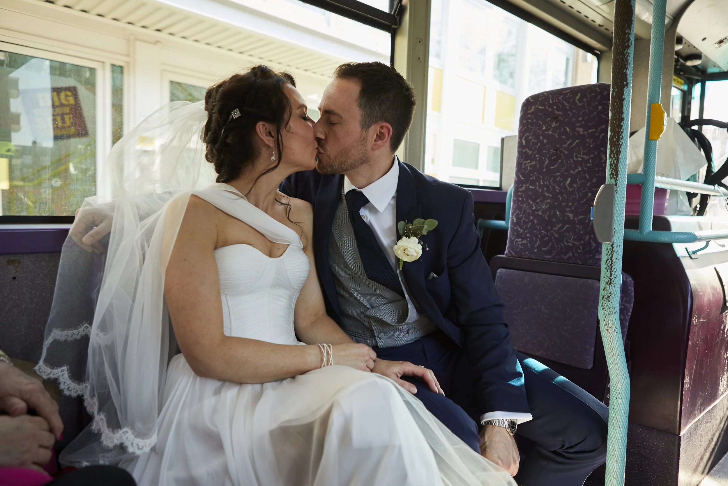 A bride and groom sharing a kiss on a bus, with the bride in a white wedding dress and veil, and the groom in a blue suit with a boutonniere, inside a bus with windows and purple seats.
